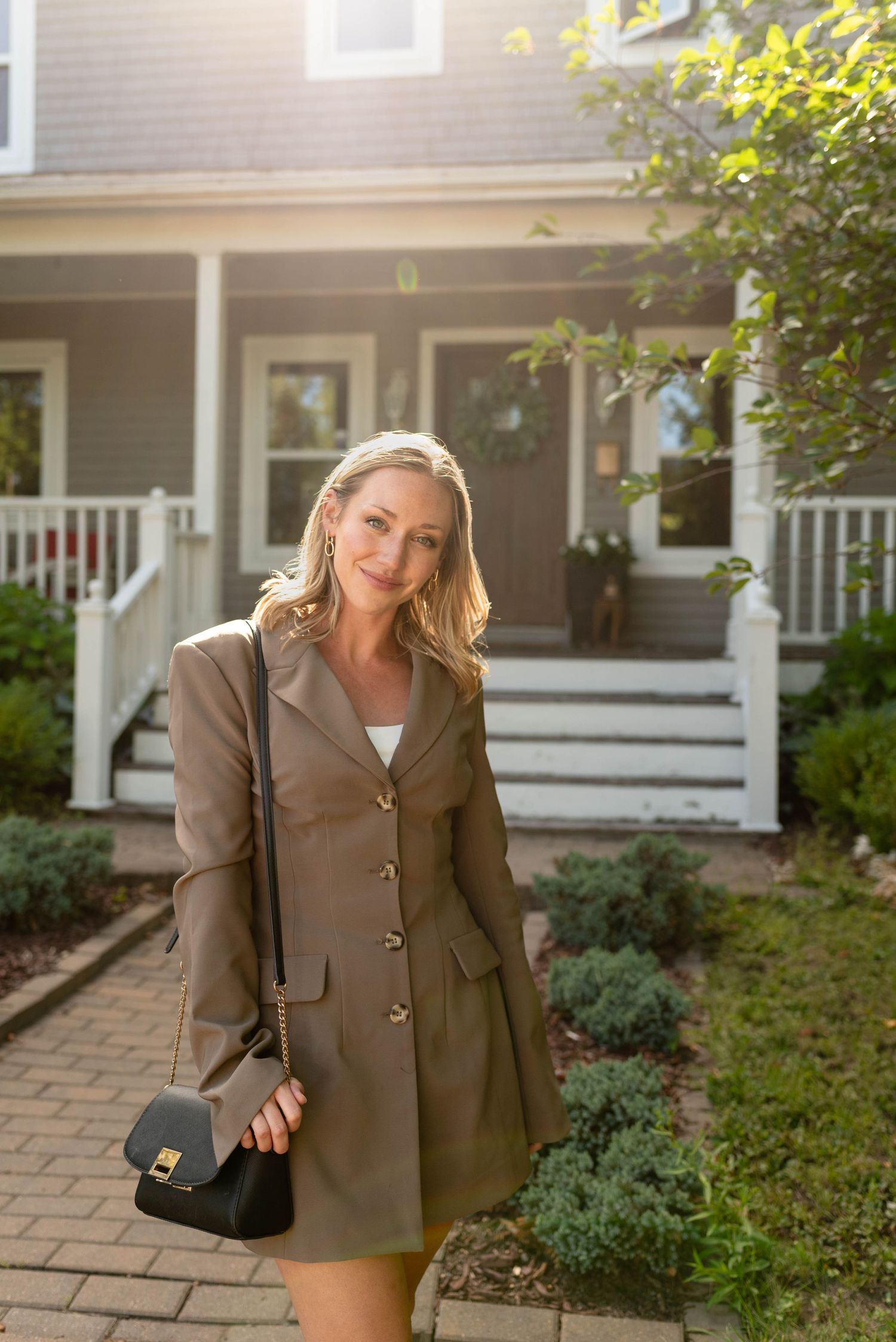 Elegant blonde woman in brown blazer dress holding black bag posing outside gray suburban house.