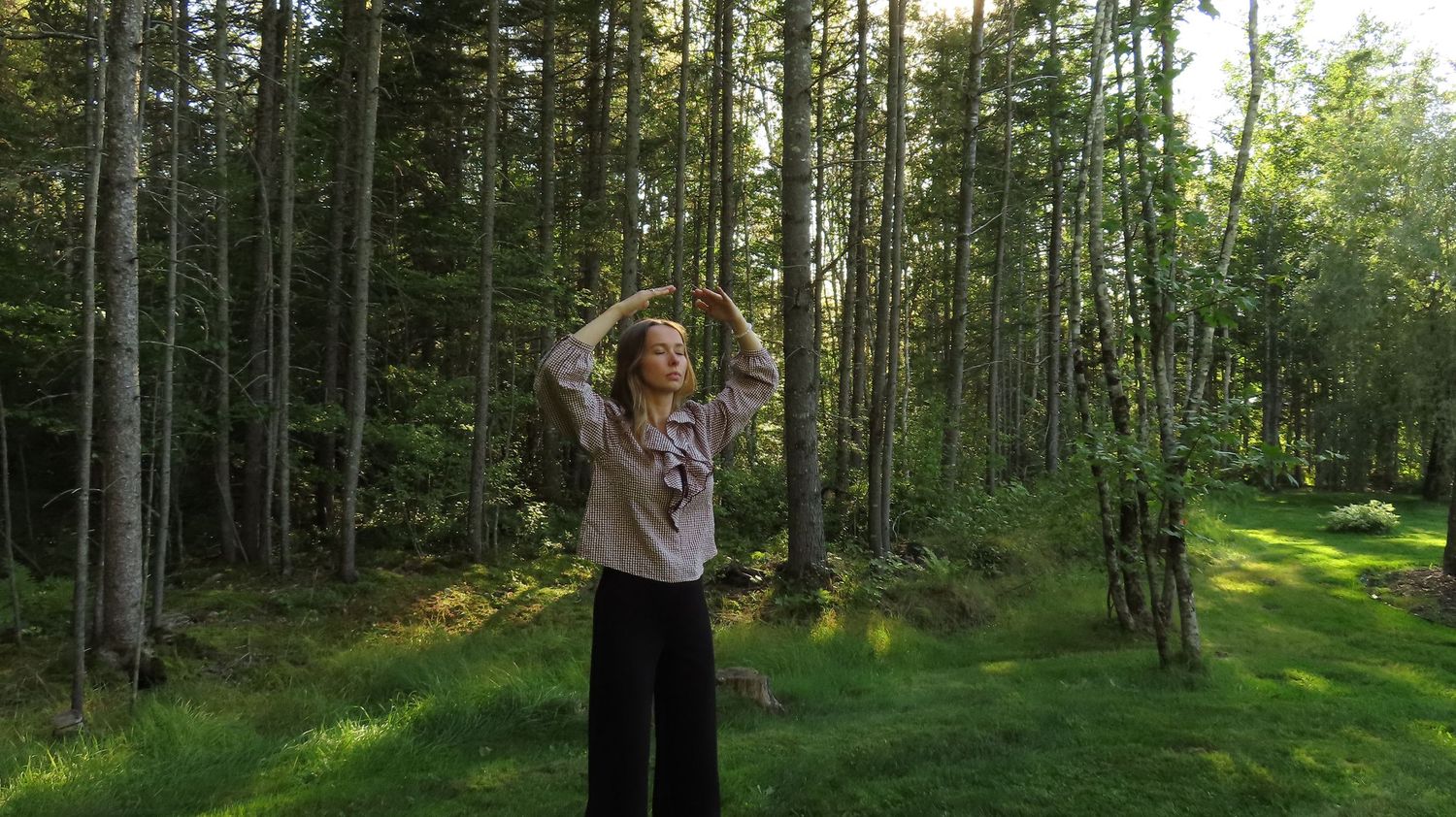 Woman standing in a sunlit forest clearing with arms raised above her head gracefully.