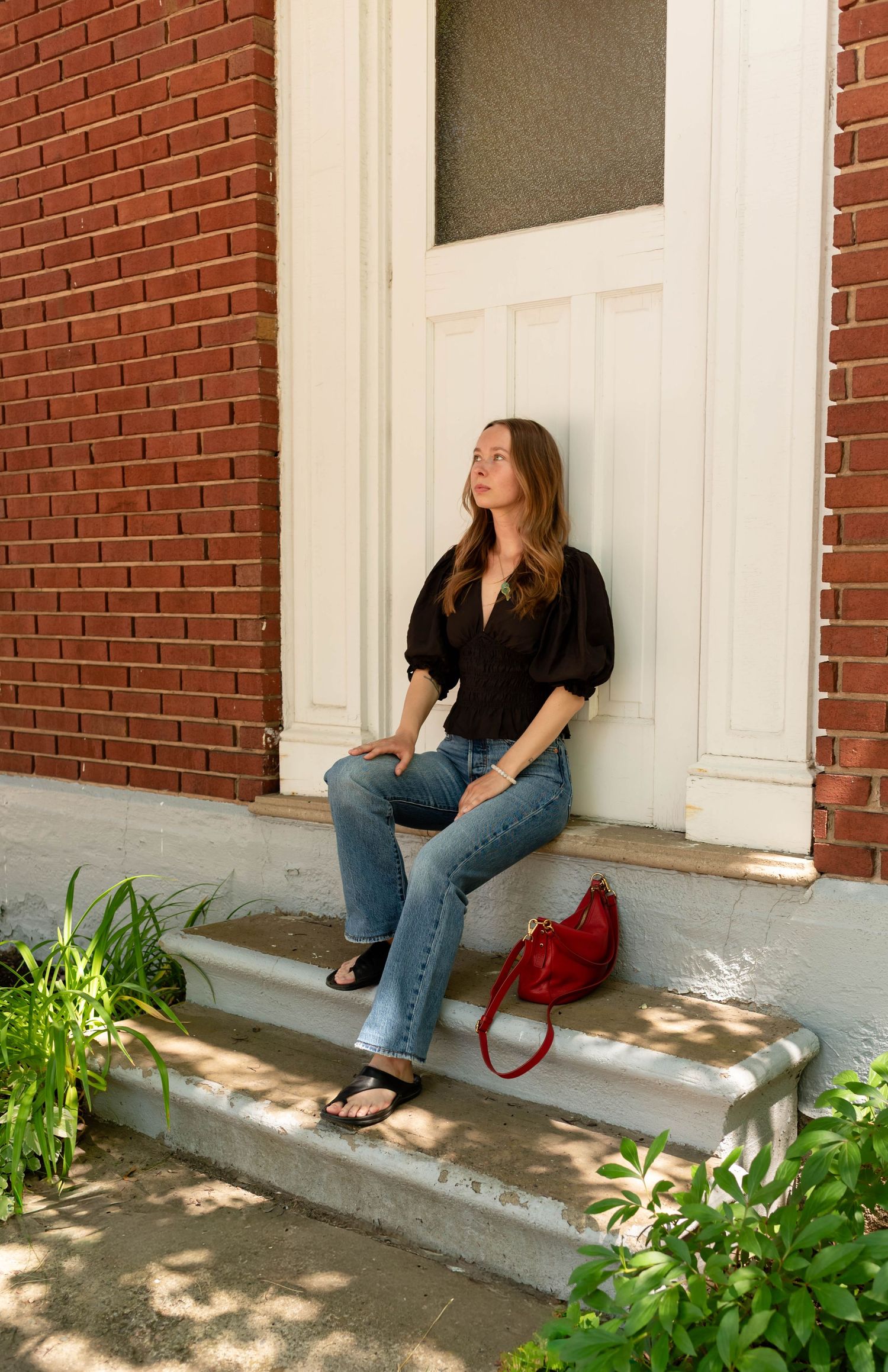 Woman in black blouse and jeans sitting on brick building steps with a red handbag.