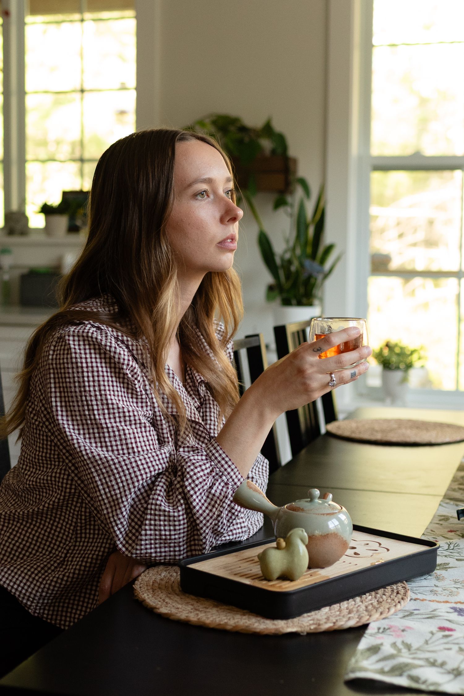 Young woman in gingham shirt holding glass teacup while gazing out window with tea set on table.