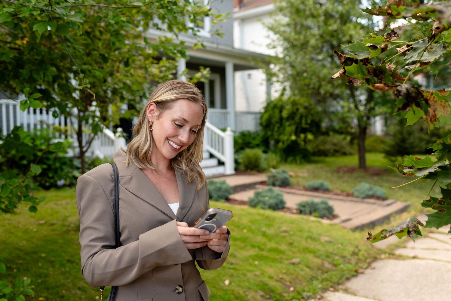 Blonde woman in beige blazer checking phone outside a house surrounded by greenery.