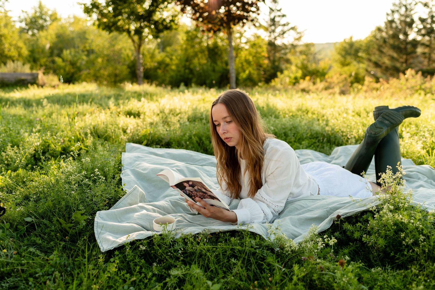 Young woman in white top and green boots lying on blanket reading book in sunny field.