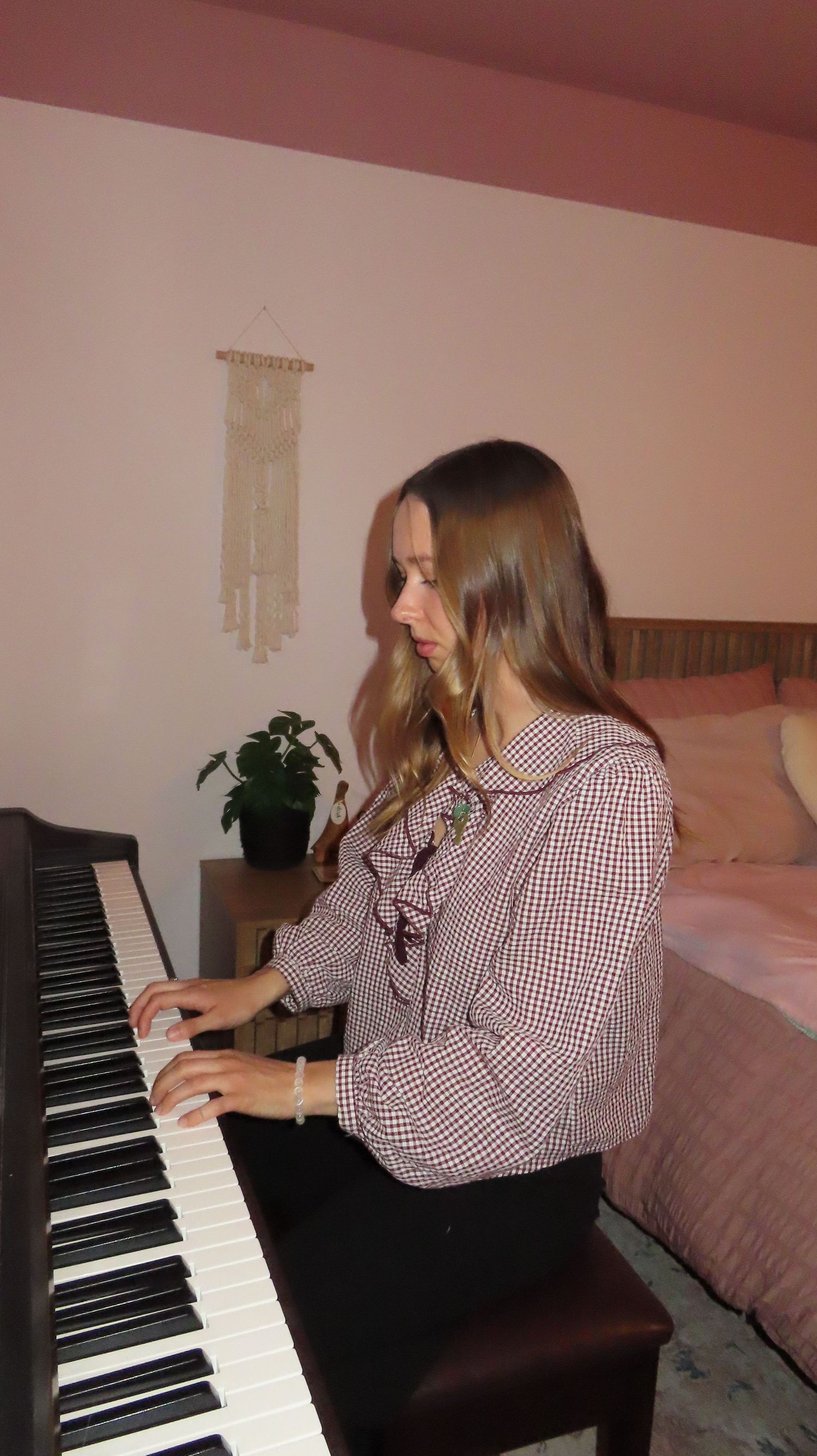 Young woman with long hair playing black piano keyboard in pink bedroom with macrame wall hanging.