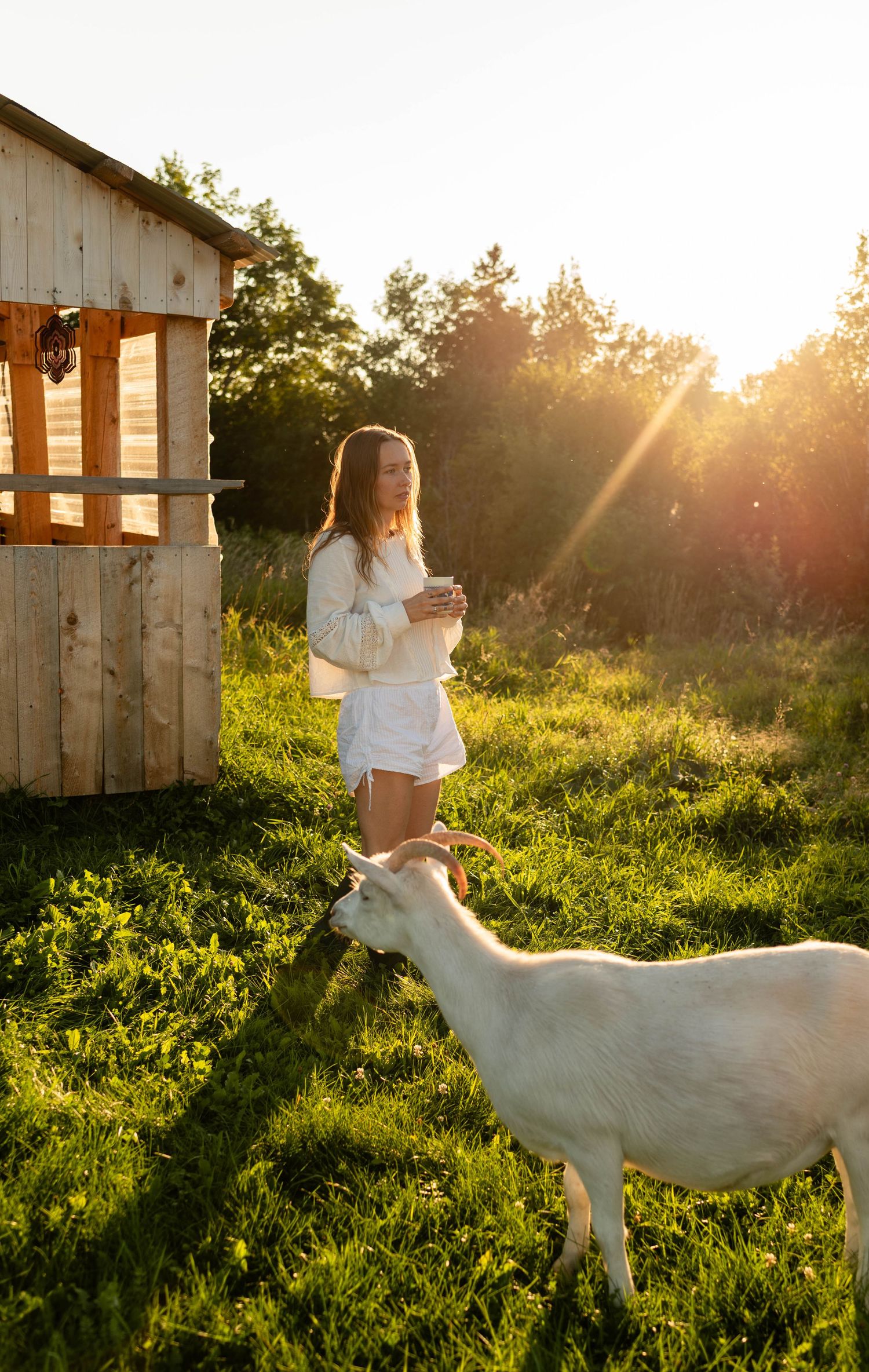 Smiling woman holding a mug while a white goat grazes beside her near a wooden structure.