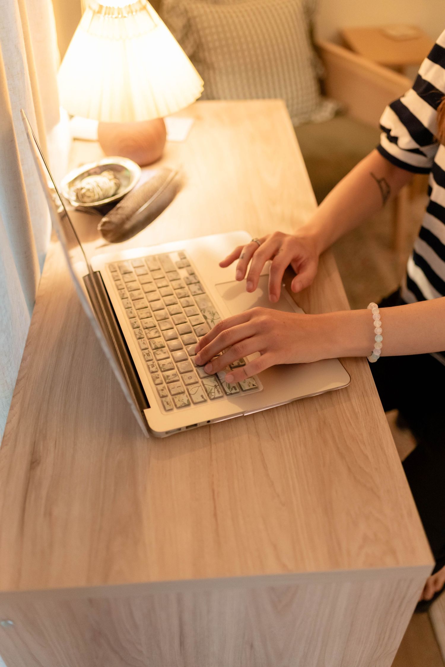 Woman typing on a gold laptop at a wooden desk with a warm lamp in a cozy room.