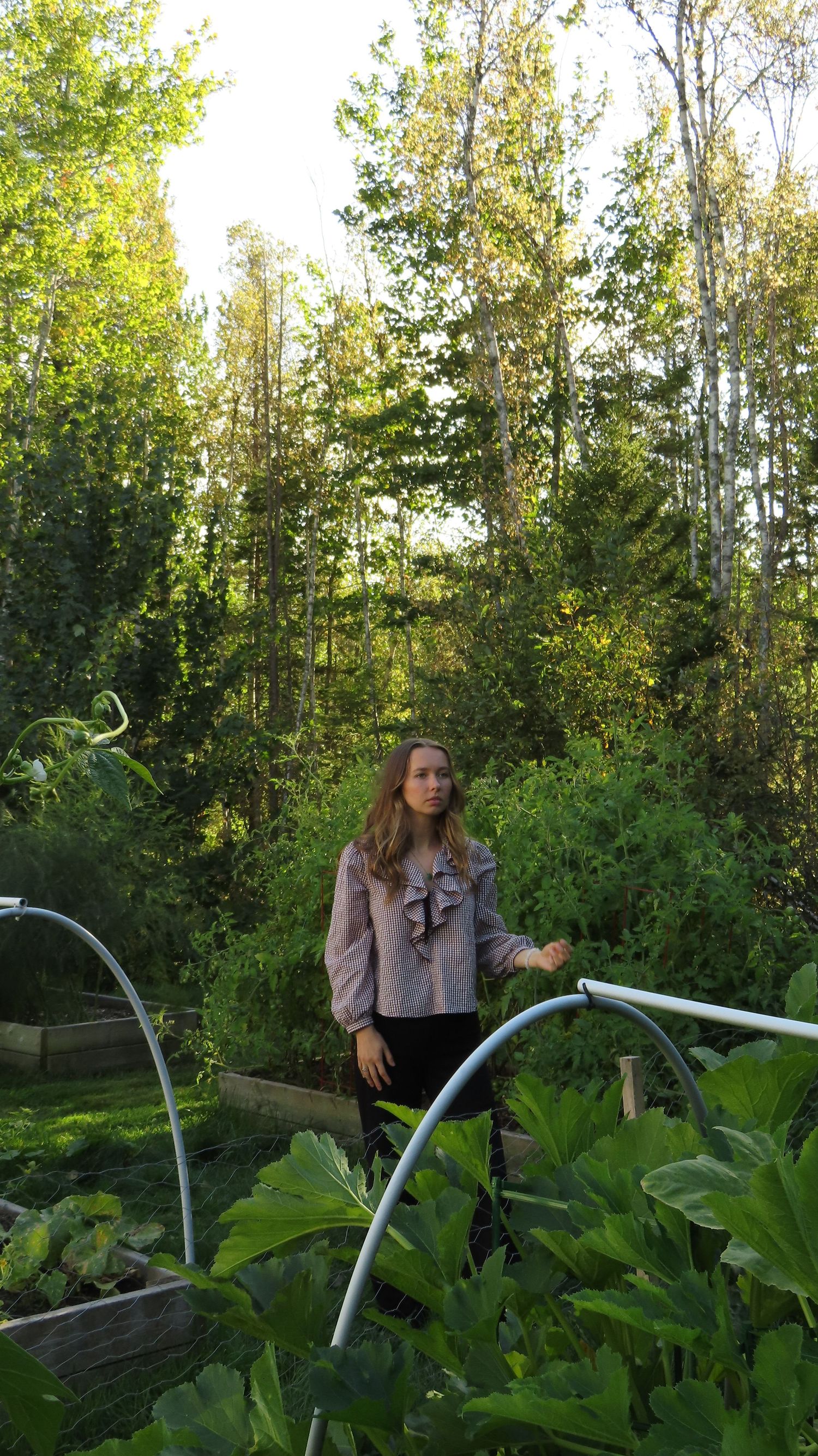 Young woman in pink ruffled blouse standing among raised garden beds with lush green trees behind.