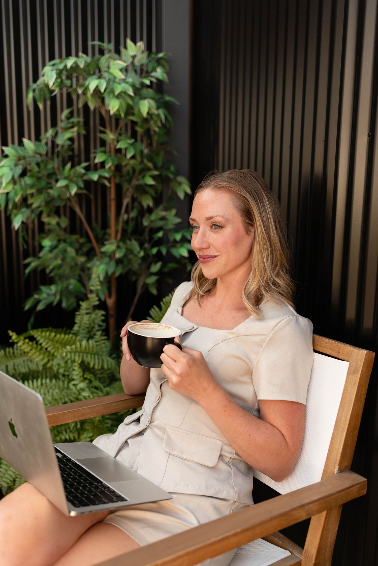 Blonde woman in beige outfit enjoying coffee with laptop in modern outdoor setting.