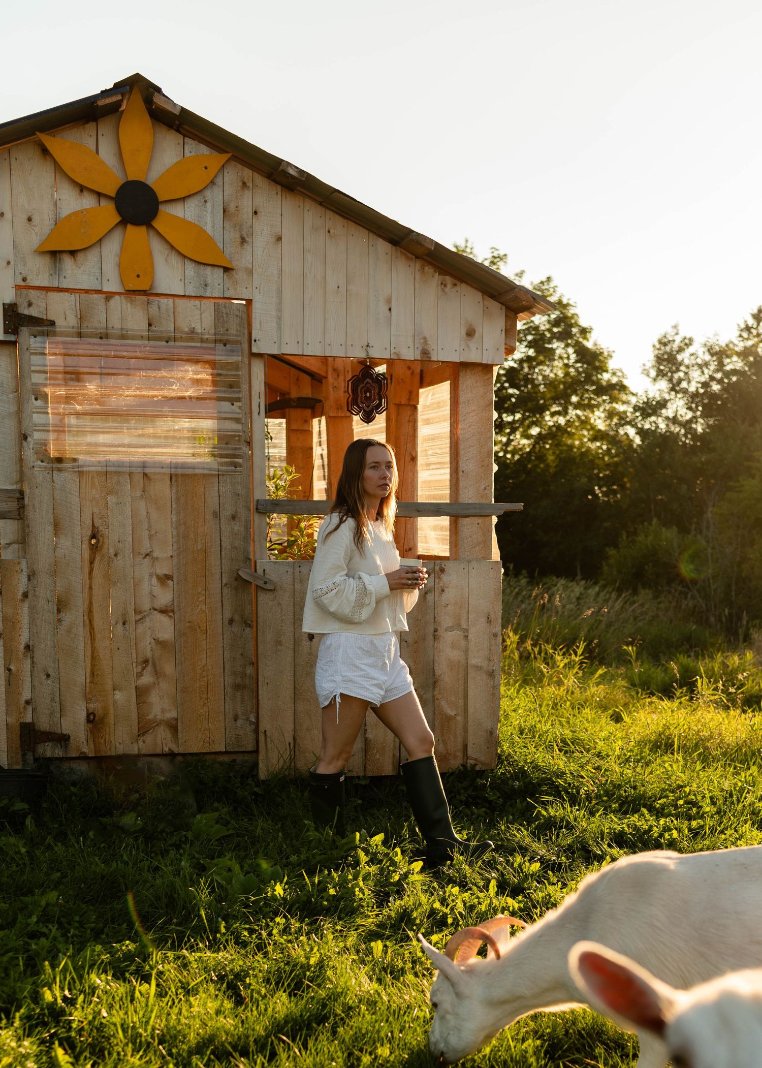 Woman in white outfit standing beside a charming wooden shed decorated with a sunflower at dusk.