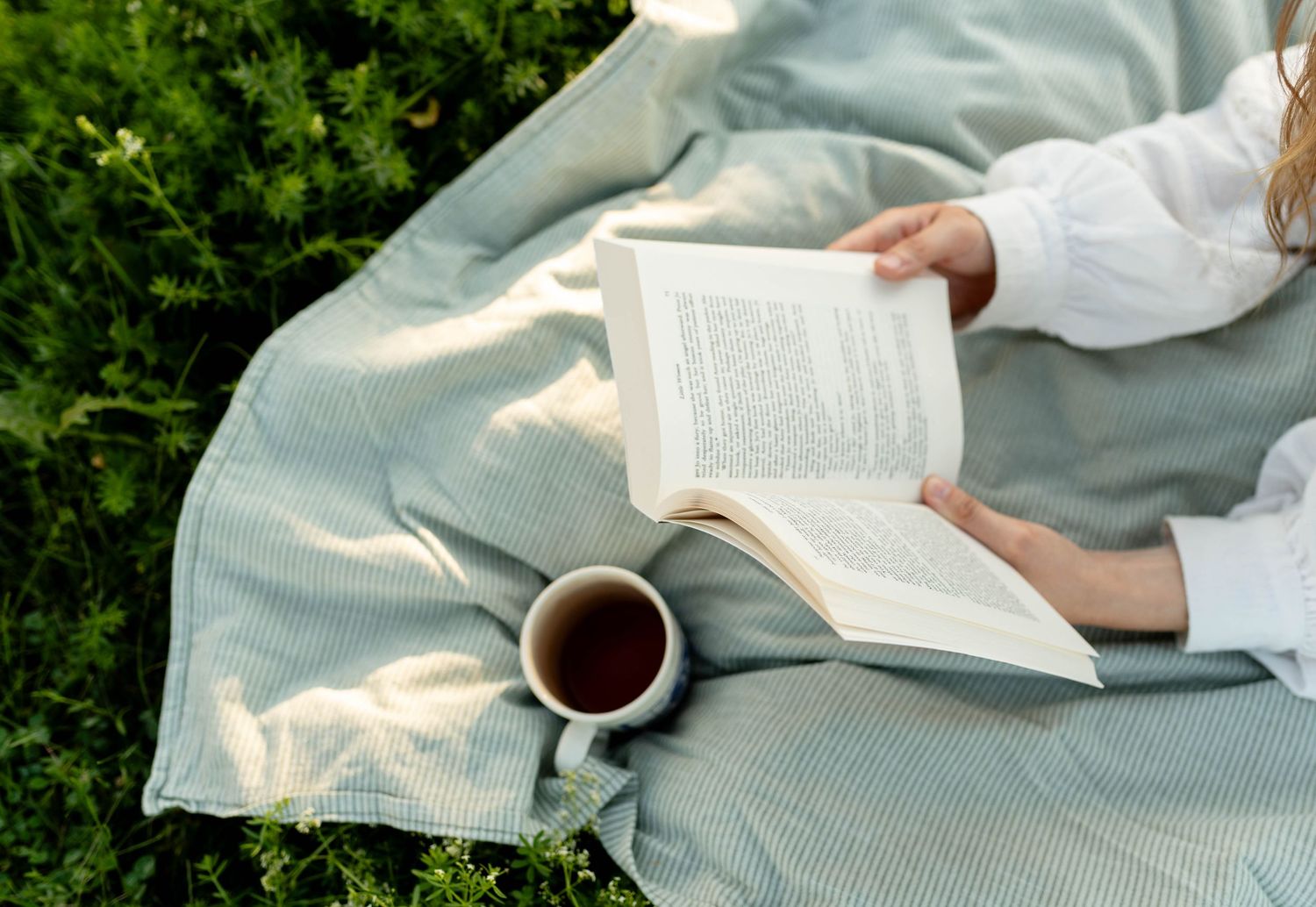 Woman in white top reading book on blanket in garden with cup of tea nearby.