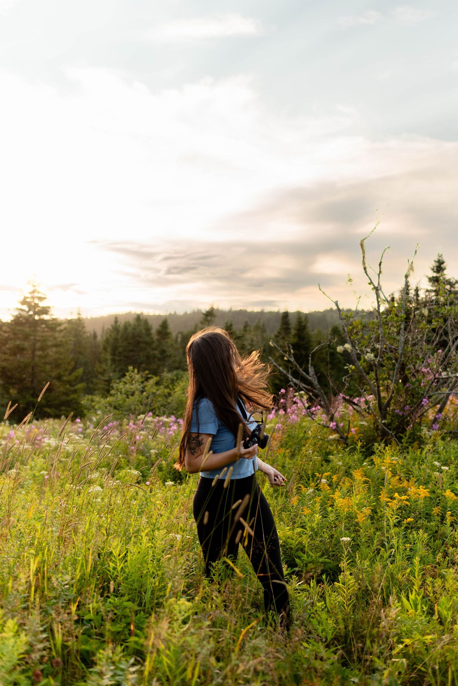 Young woman with long hair walking through a wildflower meadow at golden hour with forest backdrop.