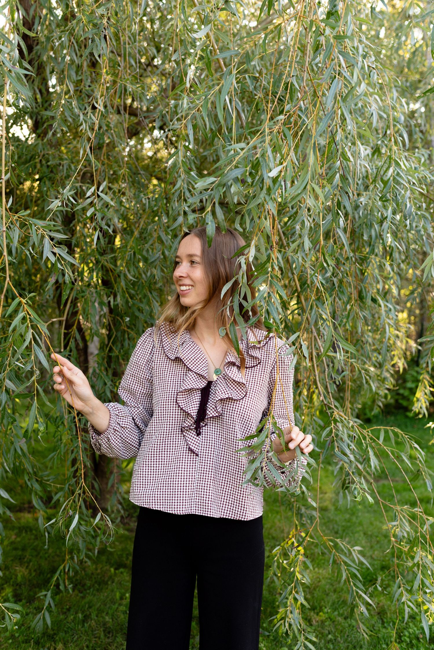 Smiling young woman in a ruffled plaid blouse holding willow branches in a lush green park.