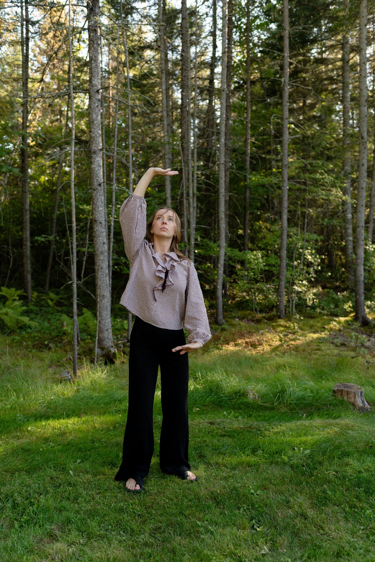 Woman in taupe blouse raising one arm upward while standing on green lawn beside pine trees.
