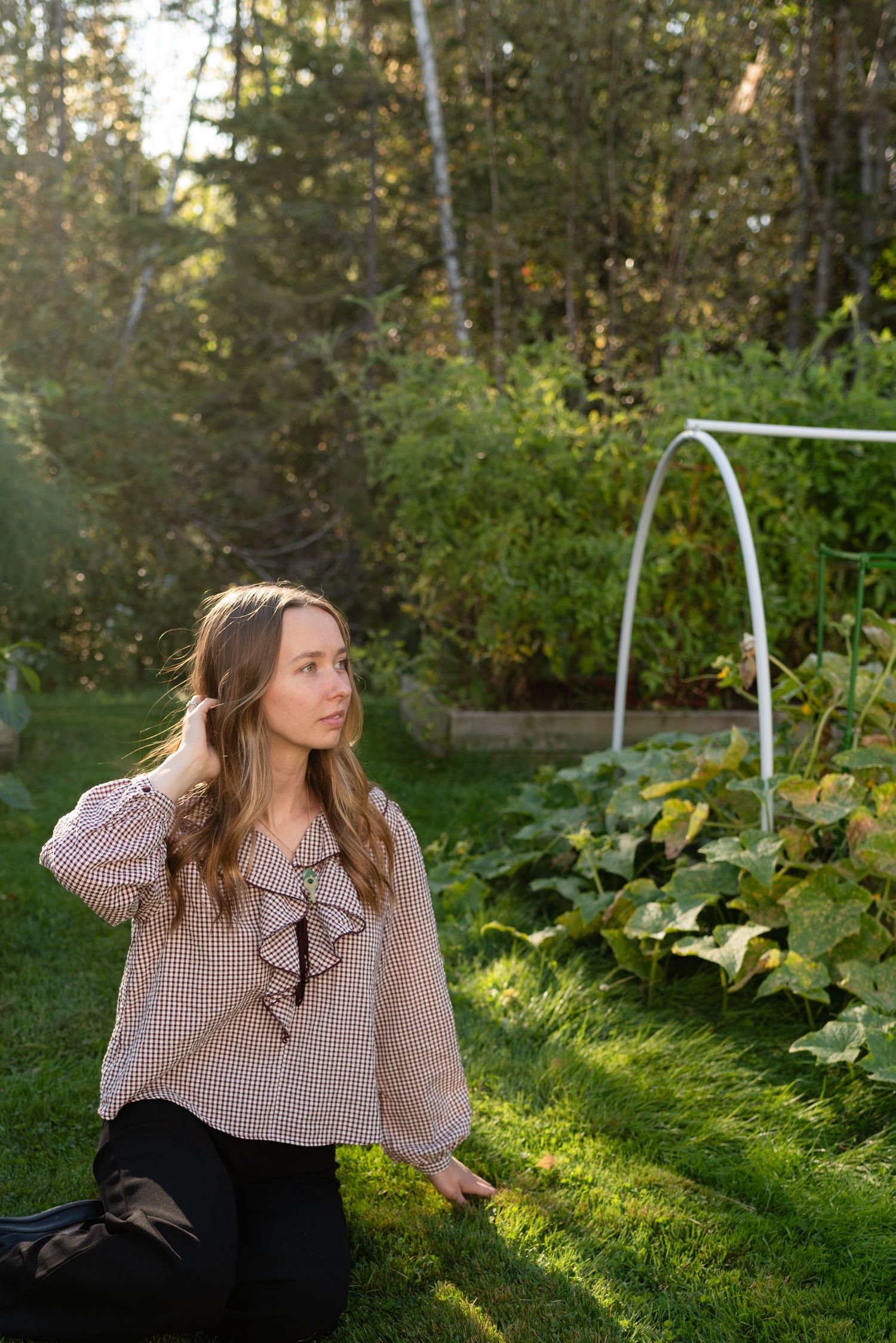 Woman in pink gingham top kneeling beside raised vegetable garden bed in golden evening sunlight.