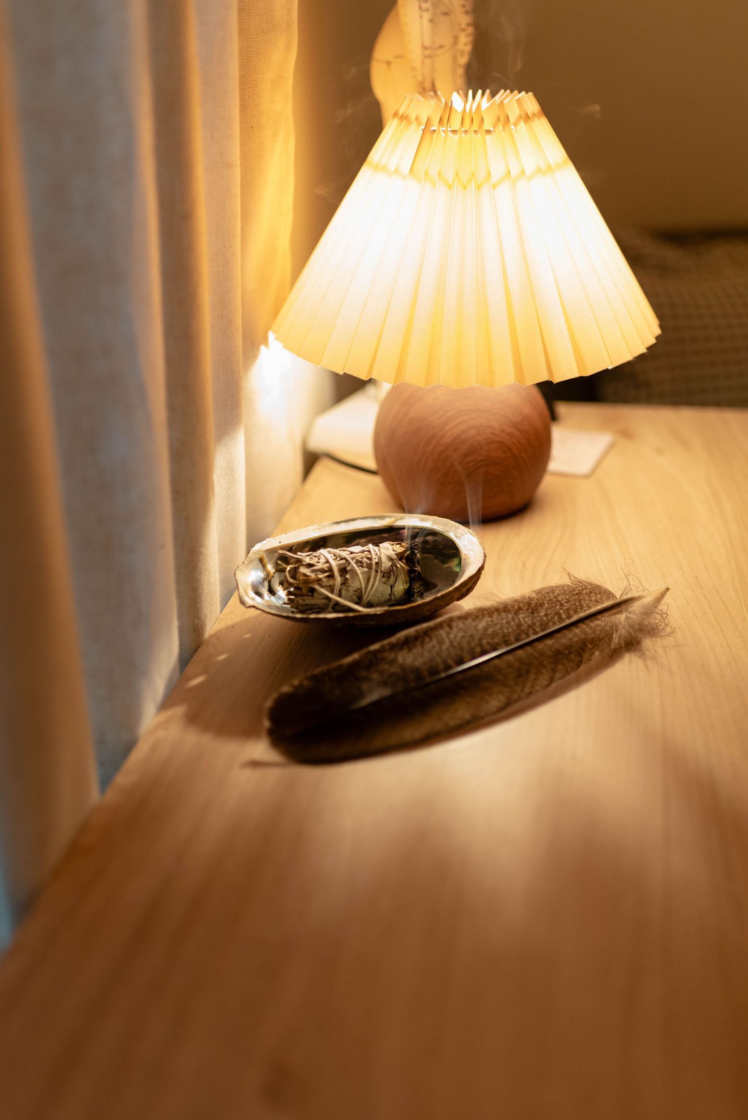 Sage smudge stick and feather resting in an abalone shell beside a warm glowing lamp.