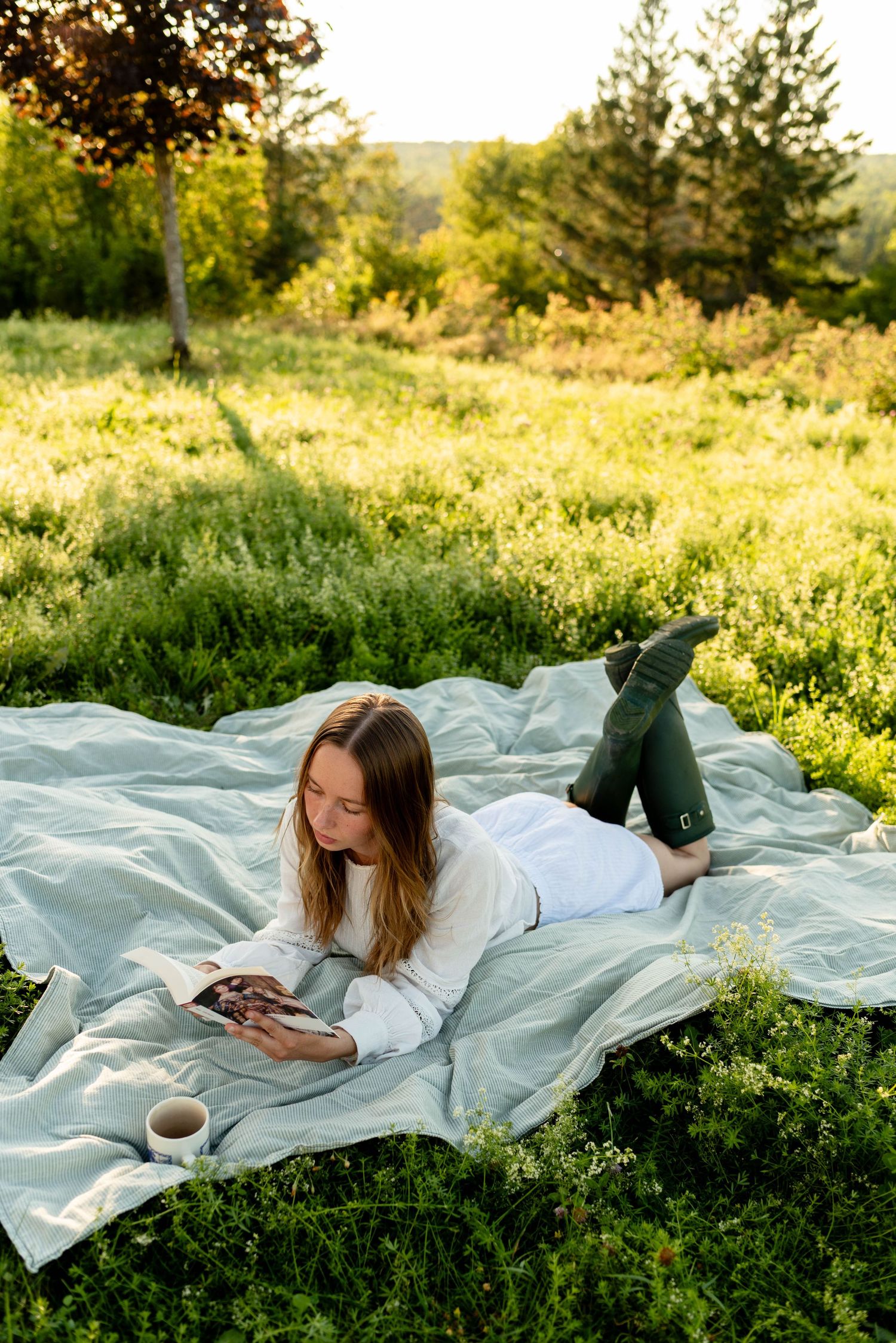 Woman in white dress and green Hunter boots lying on a blanket reading a book in a lush green field.