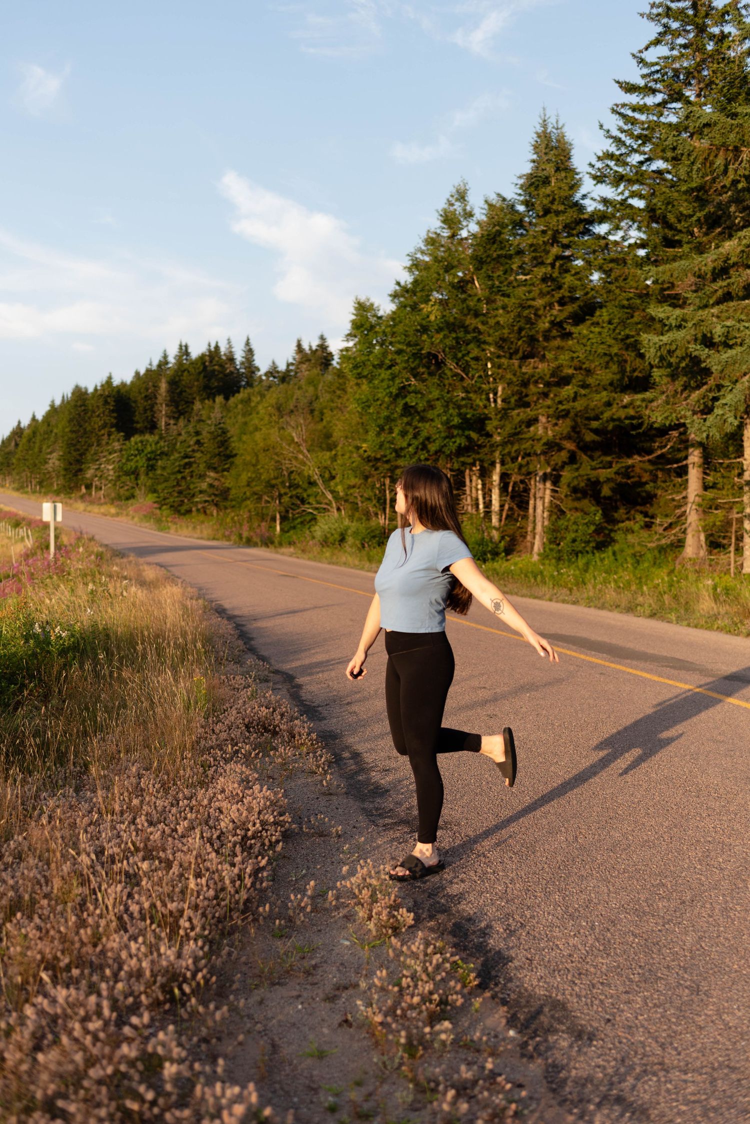 Young woman in blue top and black leggings walking along scenic forest road at golden hour.