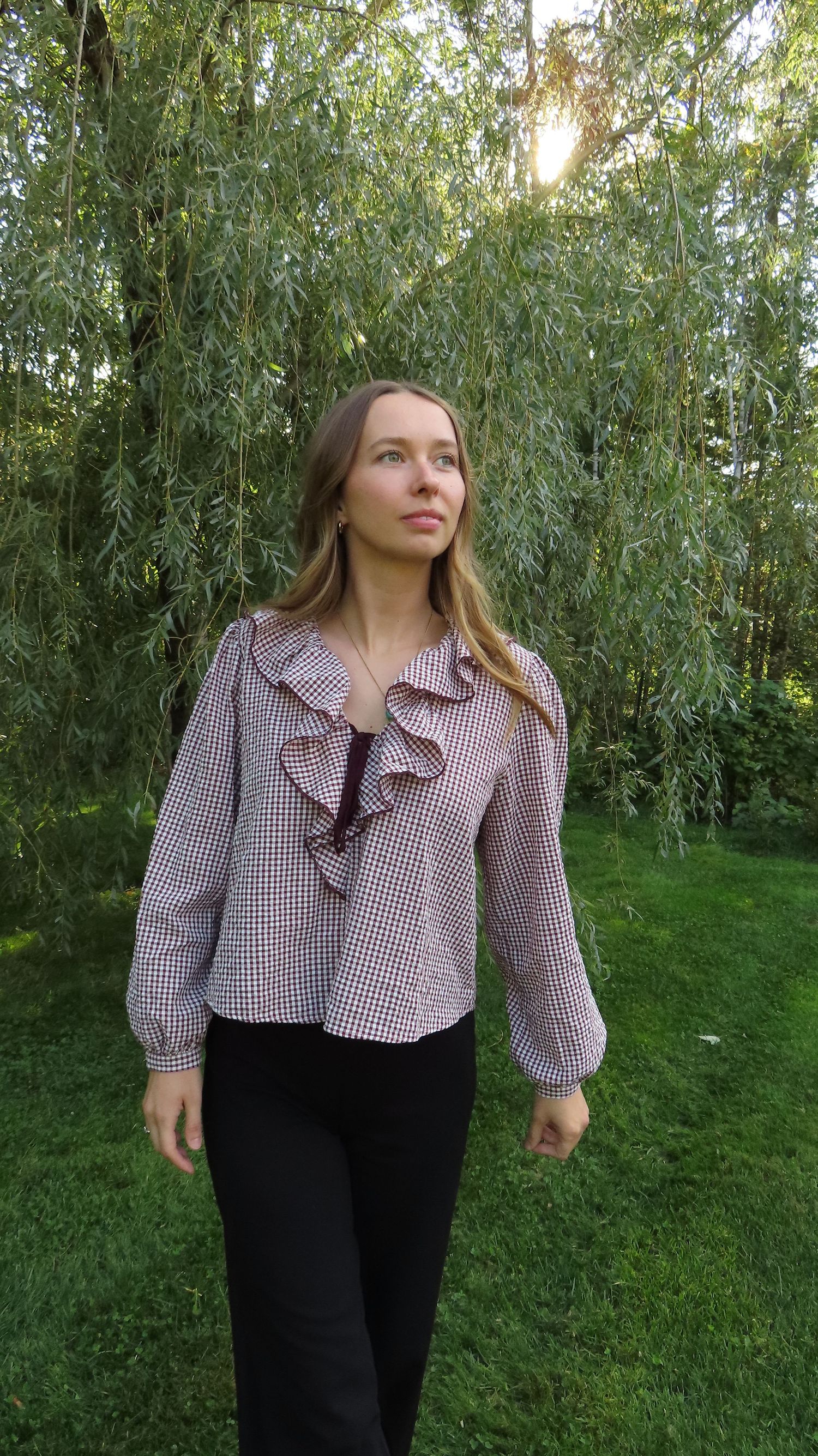 Young woman in gingham ruffle blouse and black pants standing outdoors near a weeping willow tree.