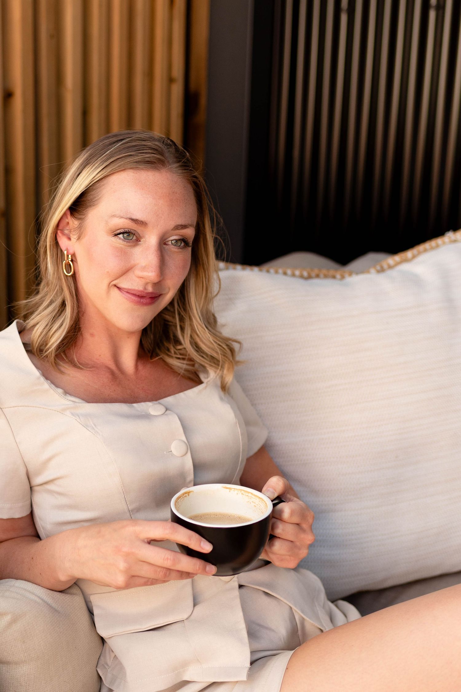 Smiling blonde woman in beige dress holding black coffee mug on cozy couch.