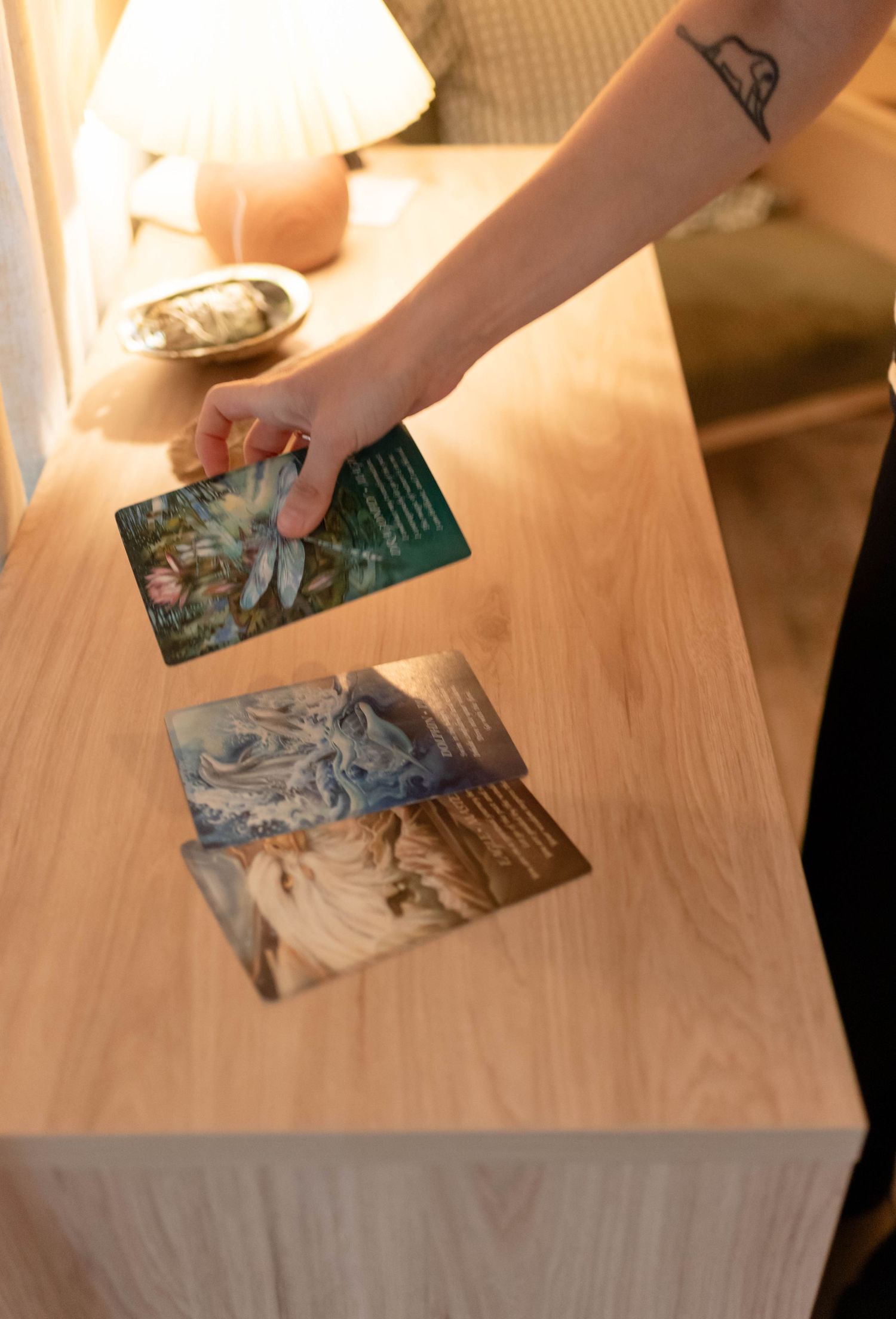 Hand with tattoo placing oracle tarot cards on wooden table in warmly lit spiritual wellness space.