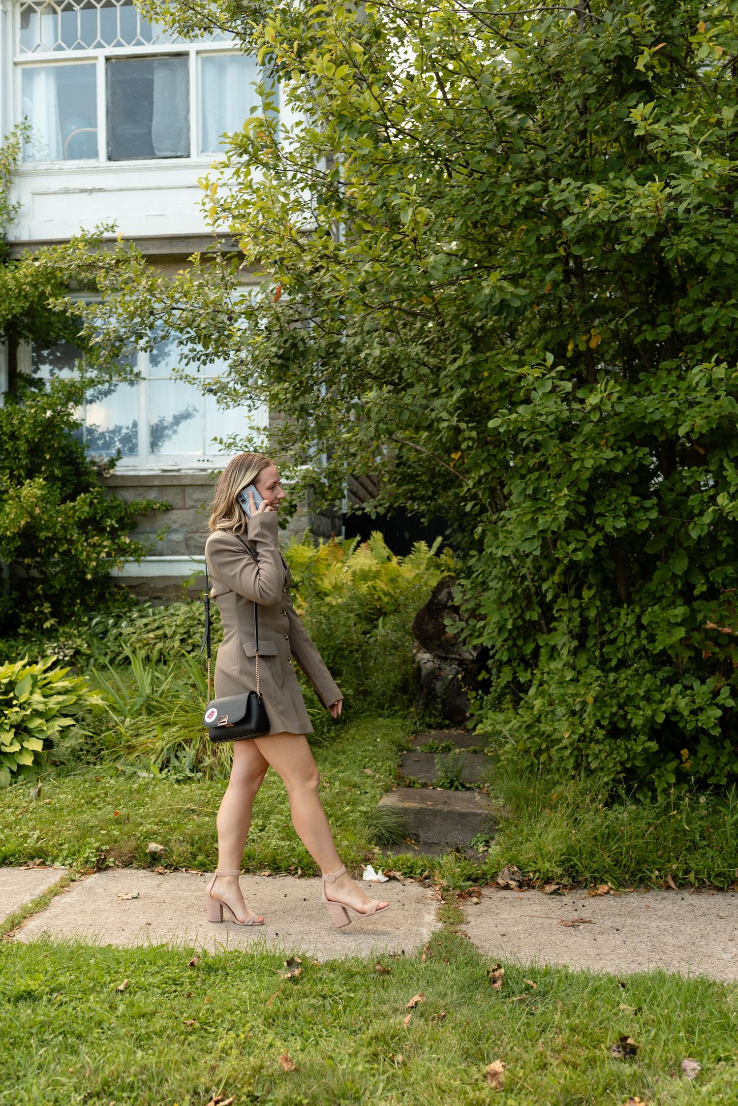 Blonde woman in taupe blazer dress walking on sidewalk while talking on phone outdoors.