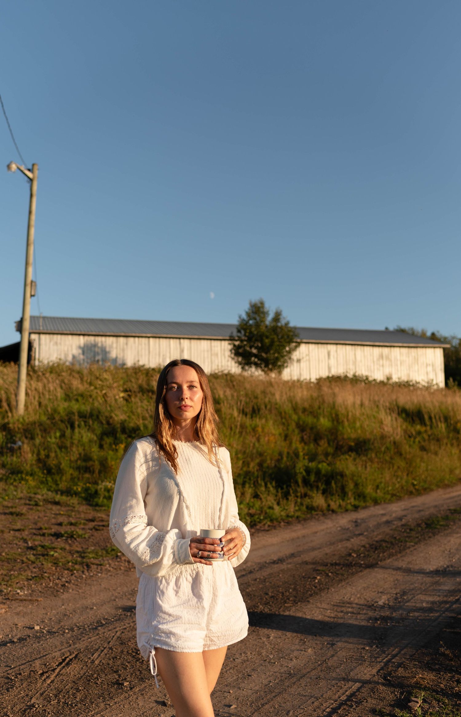 Young woman in white shorts set holding a cup on a rural dirt road at golden hour.
