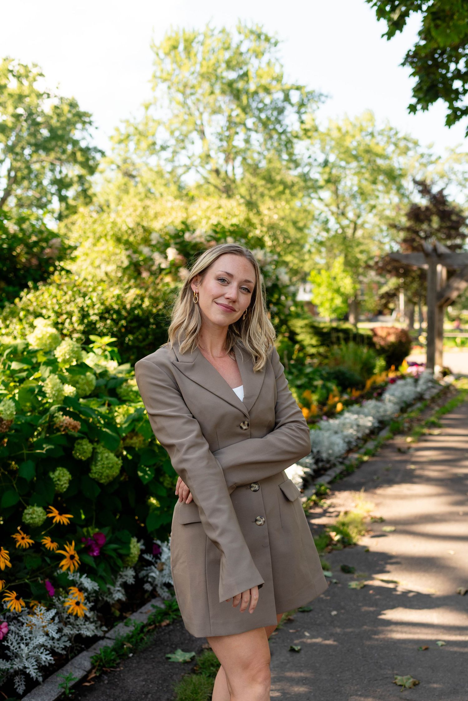 Blonde woman in khaki blazer dress posing with arms crossed beside a colorful garden.