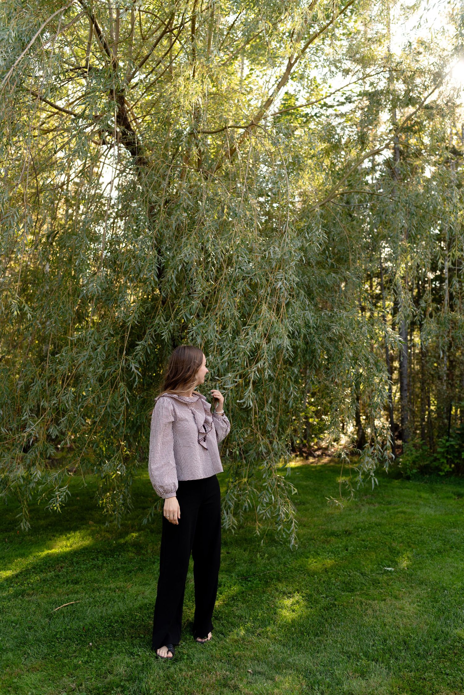 Woman in lavender blouse and black pants standing under weeping willow tree in lush green garden.
