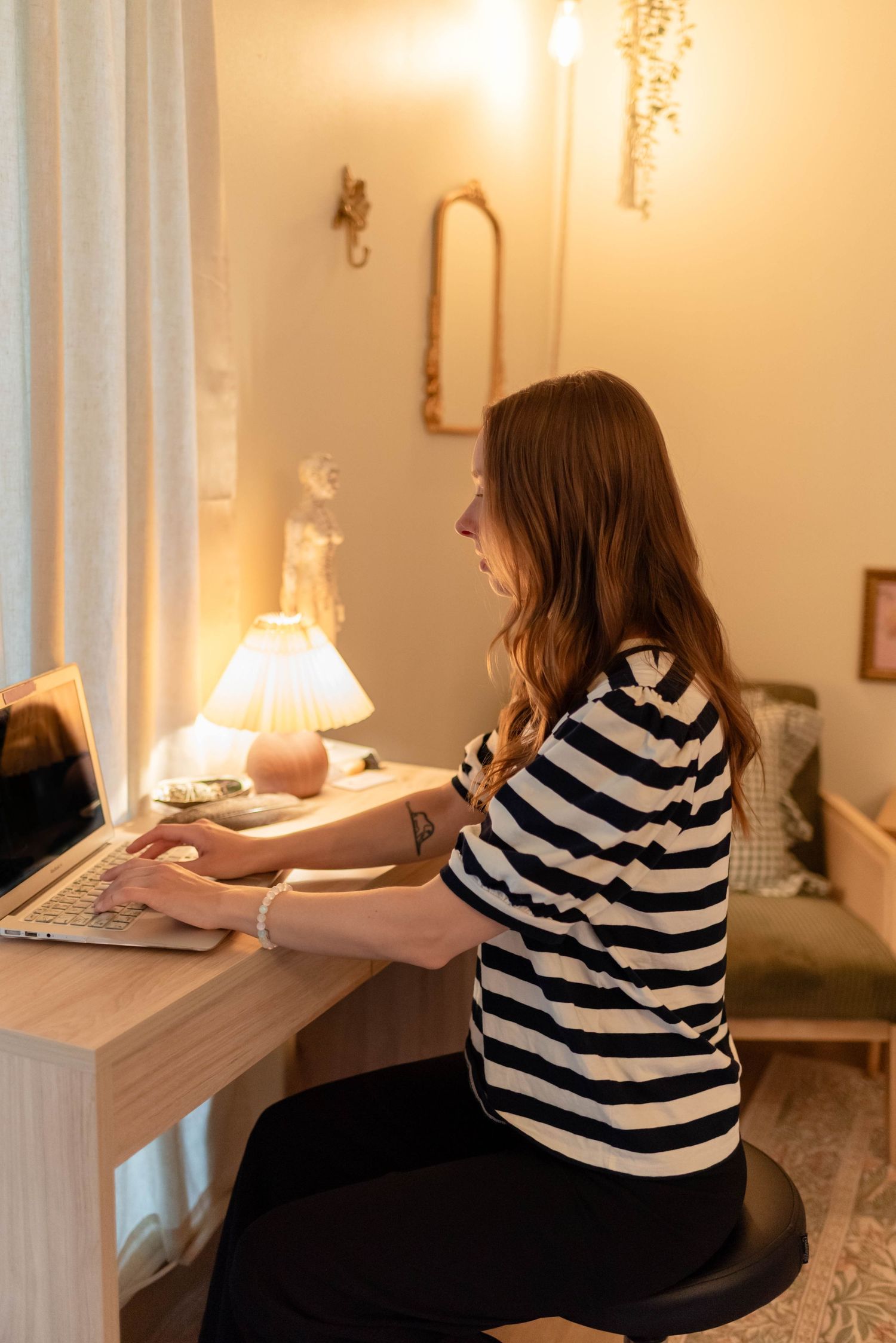 Young woman in striped shirt working on laptop at a cozy warmly lit wooden desk.