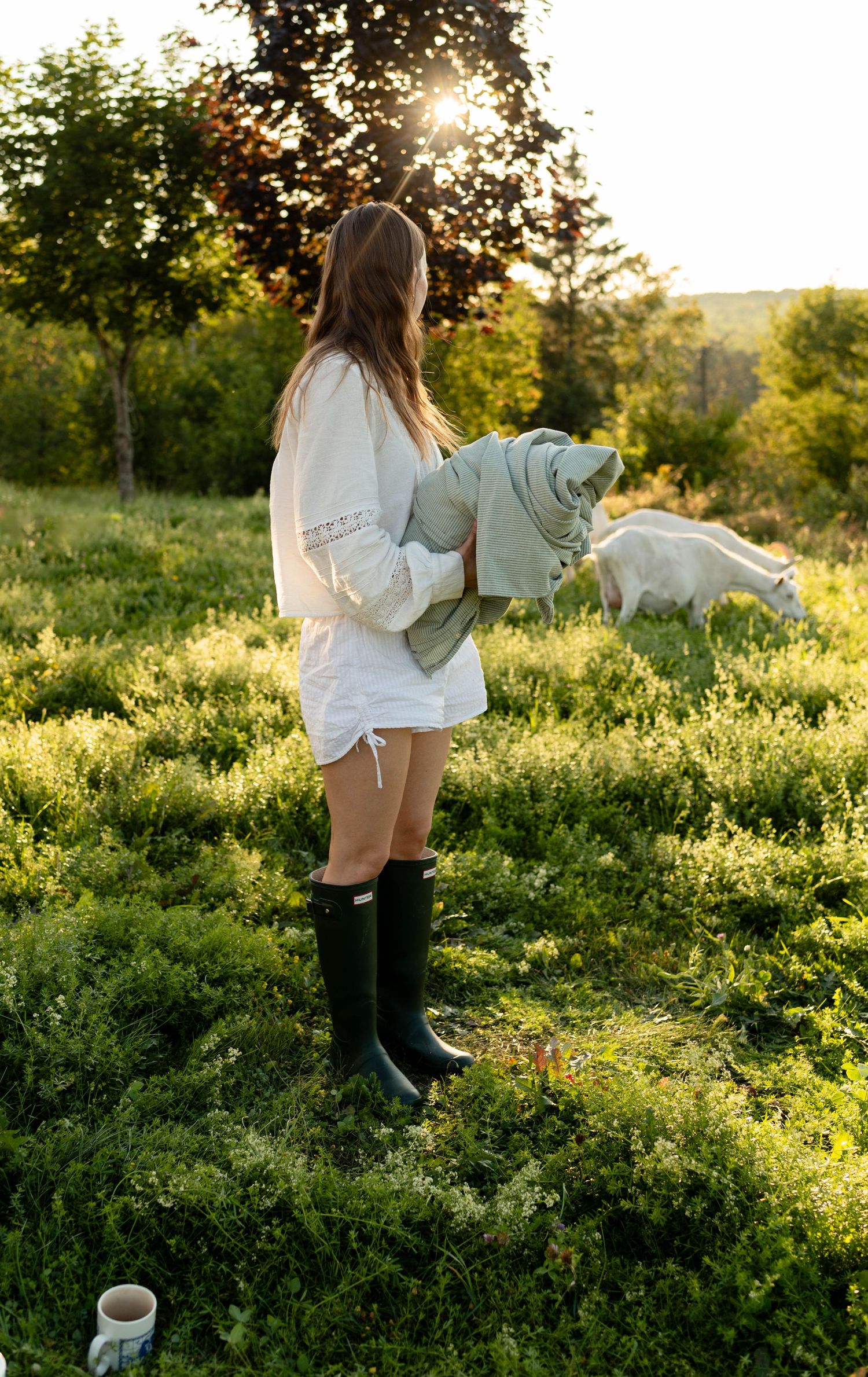 Woman in white dress and green Hunter boots holding a blanket in a sunlit field with grazing goats.