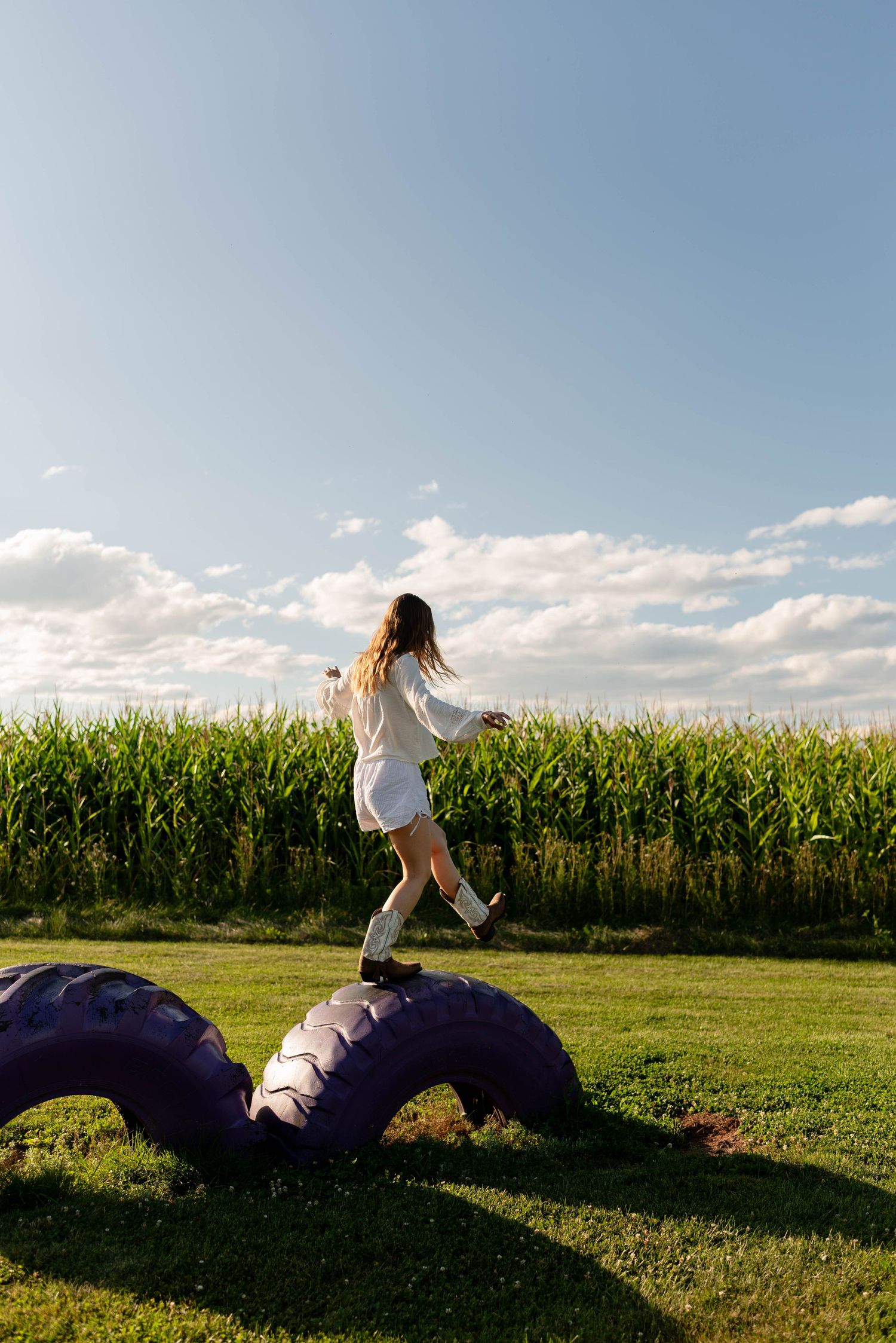 Woman in white dress balancing on large tires in a sunny green cornfield landscape.