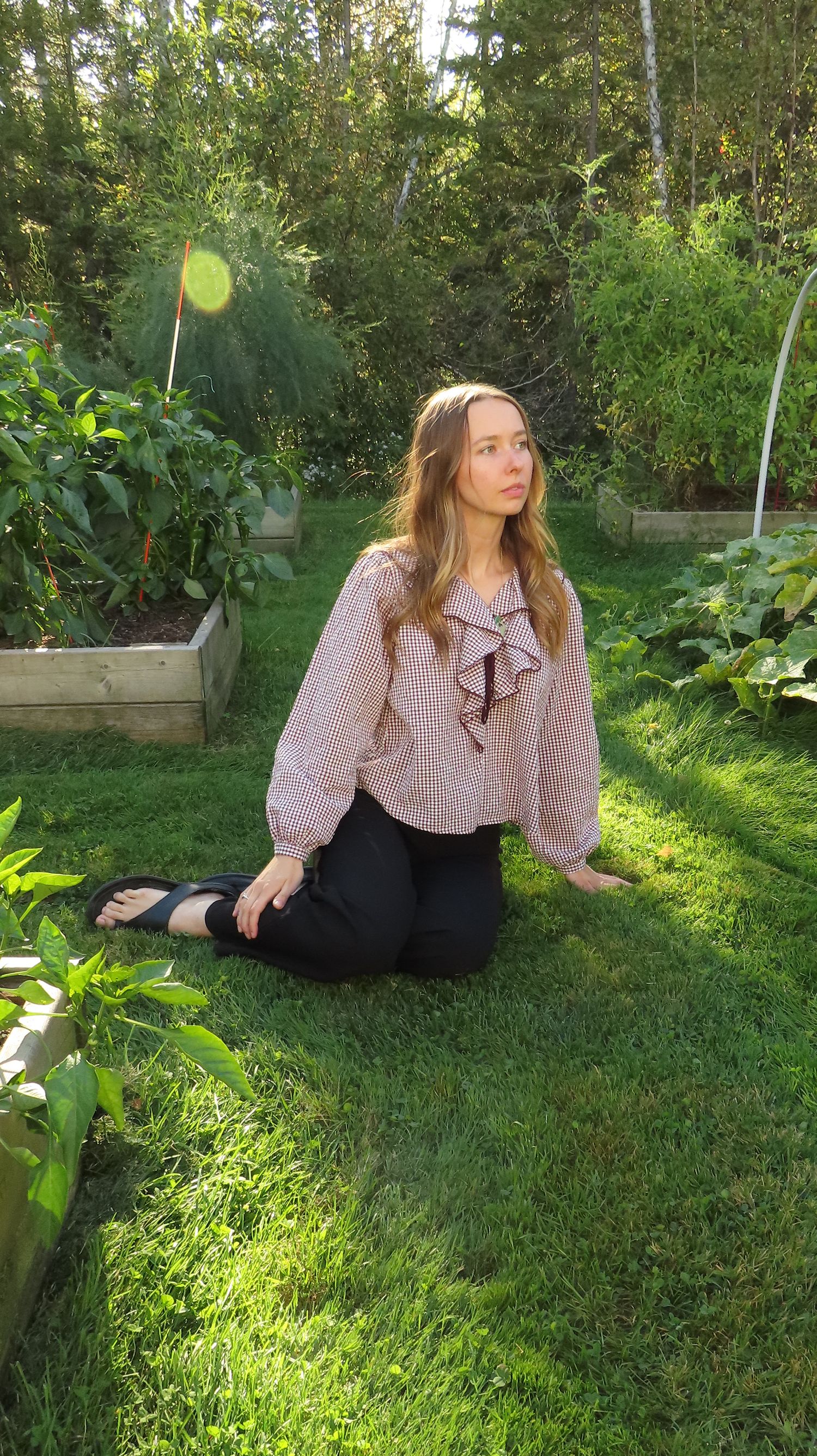 Young woman in pink gingham ruffle blouse sitting on grass in a sunny vegetable garden.