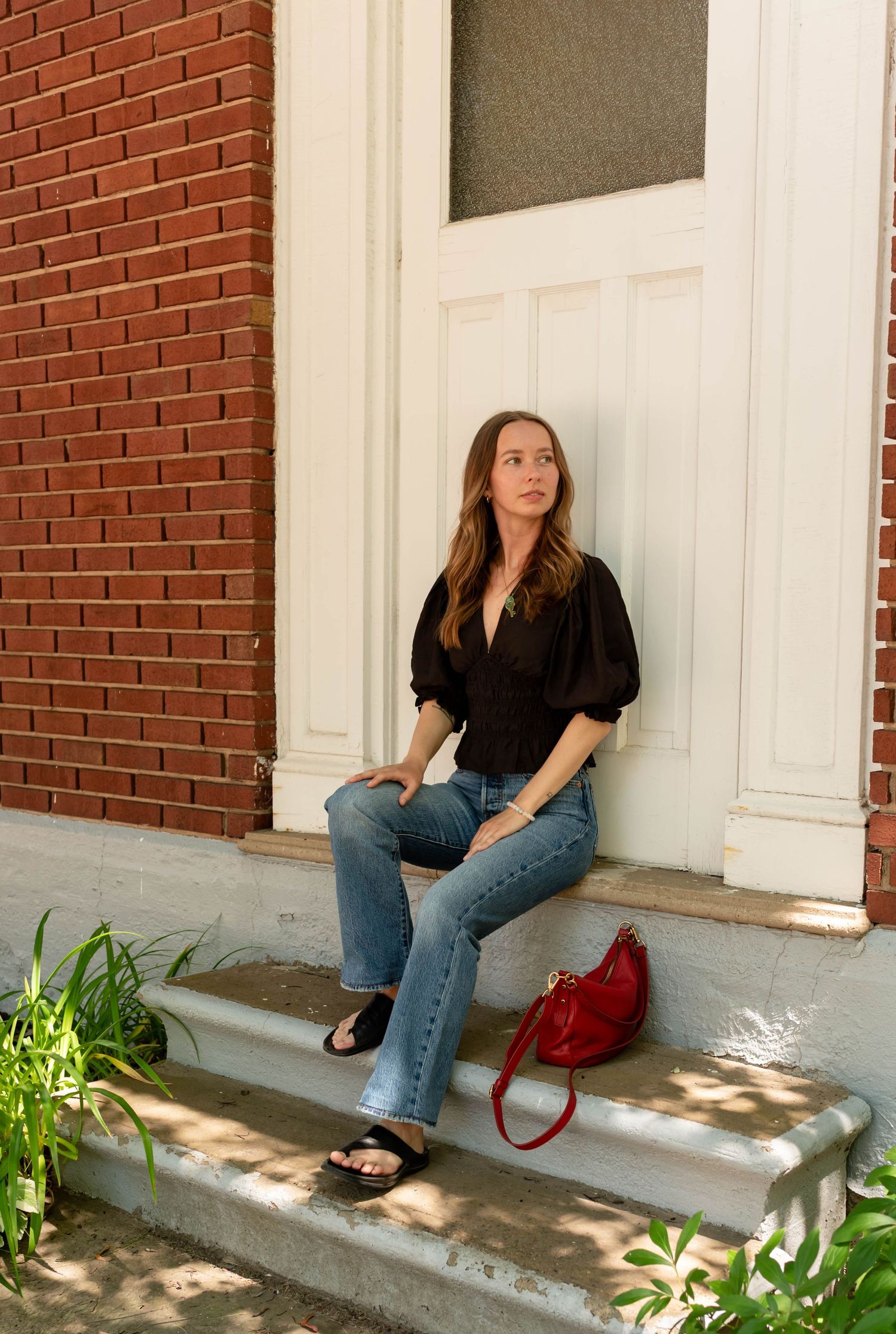 Young woman in black blouse and jeans sitting on brick building steps with red handbag.