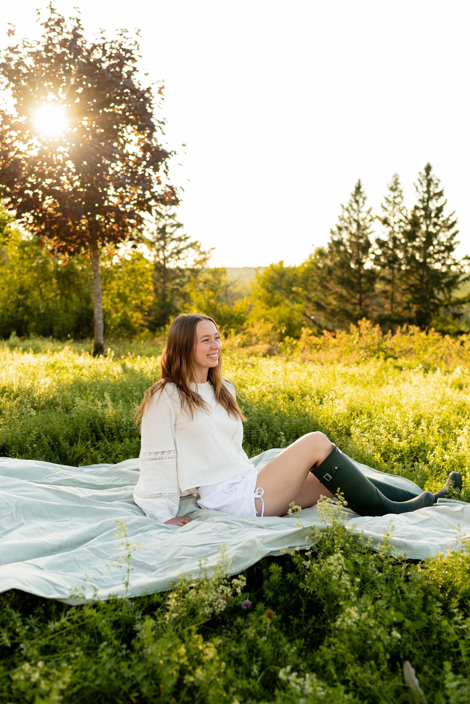 Smiling woman in white dress and green rain boots relaxing on blanket in sunny meadow.