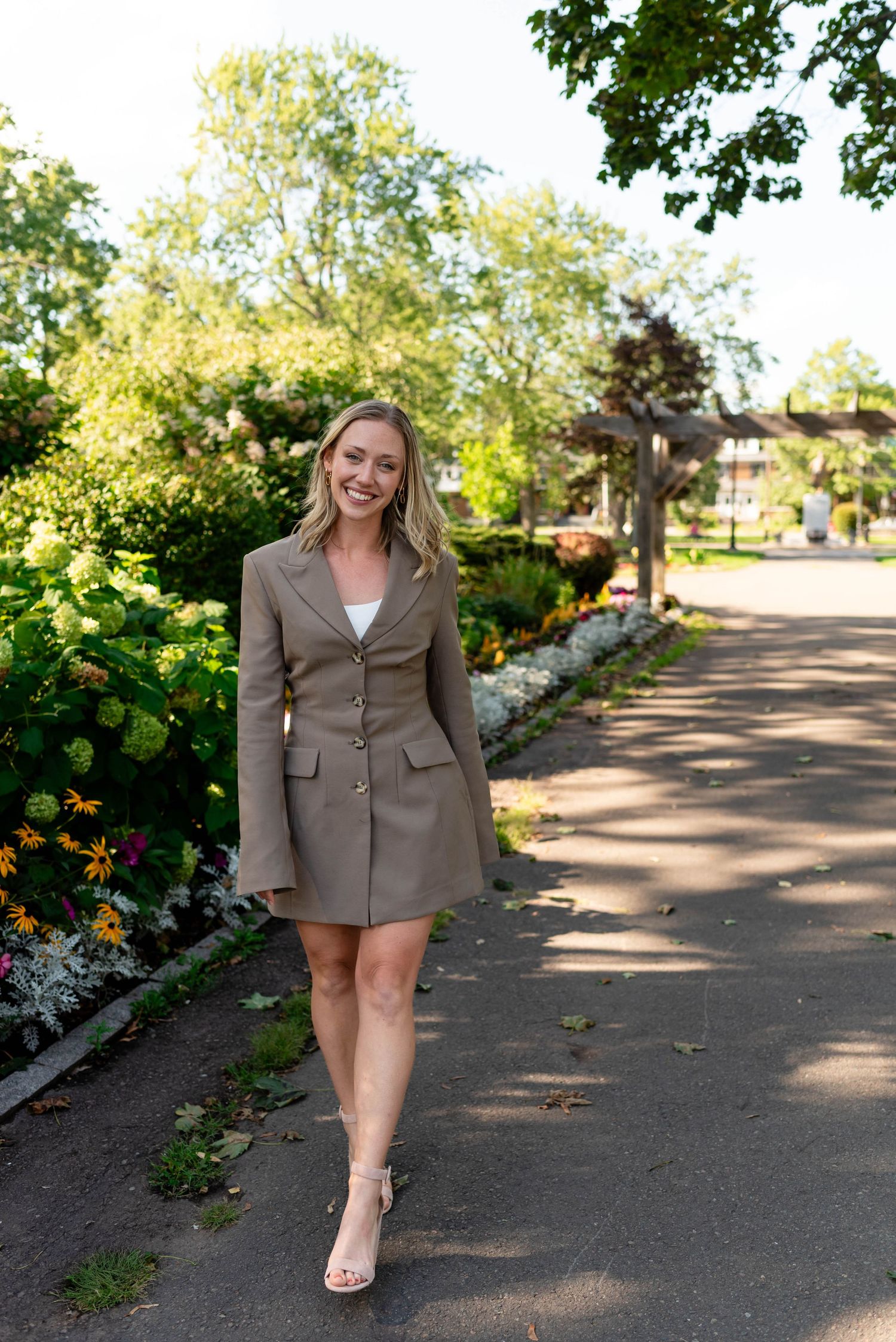 Blonde woman in taupe blazer dress walking along a flower-lined garden path in heels.