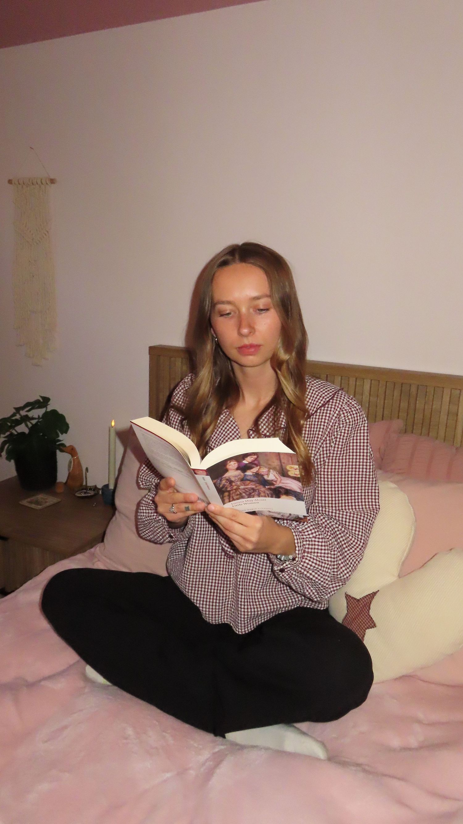 Woman in checkered shirt reading a book while sitting cross-legged on a pink bedspread.