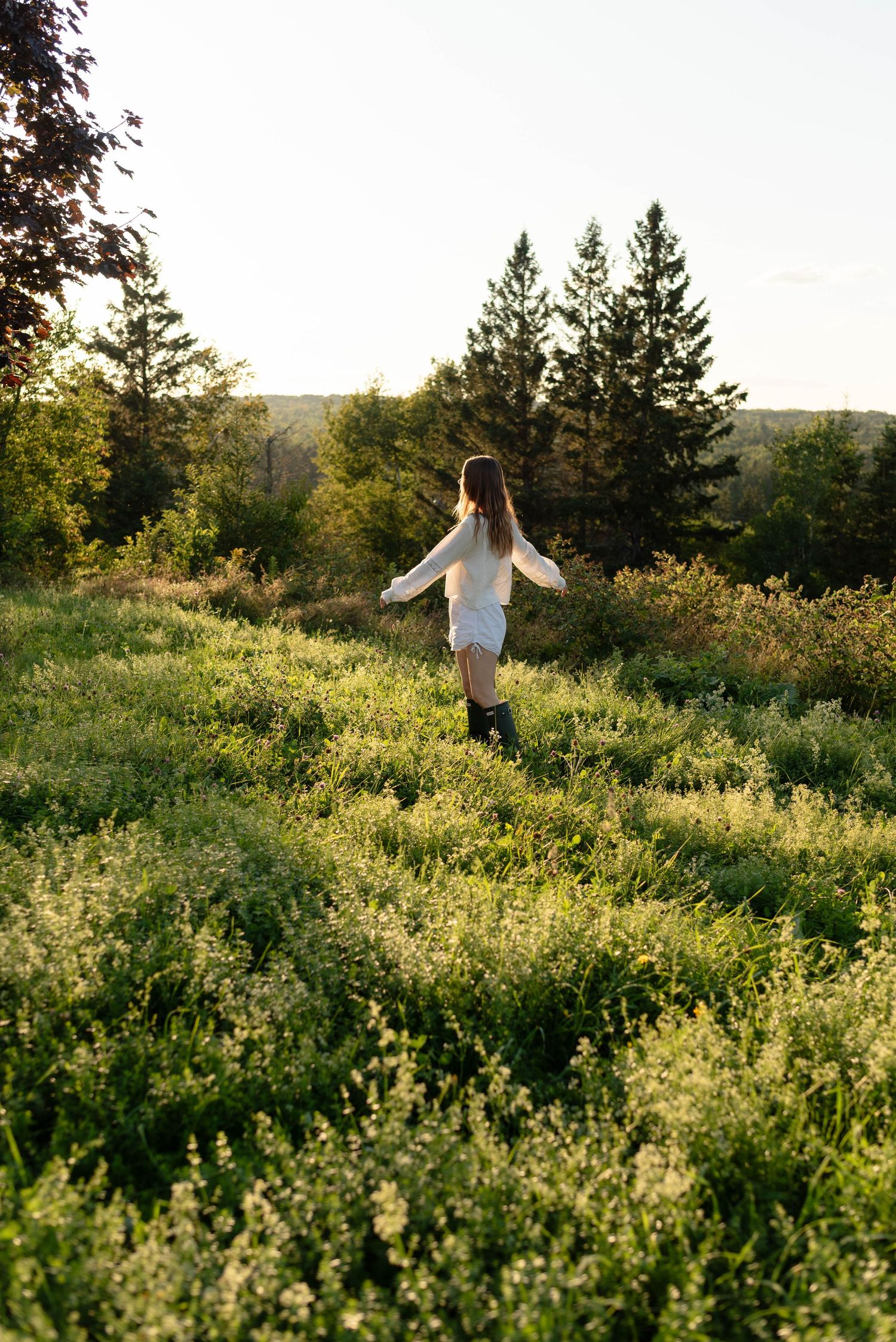 Woman in white dress and black boots walking through a sunlit green field with trees.