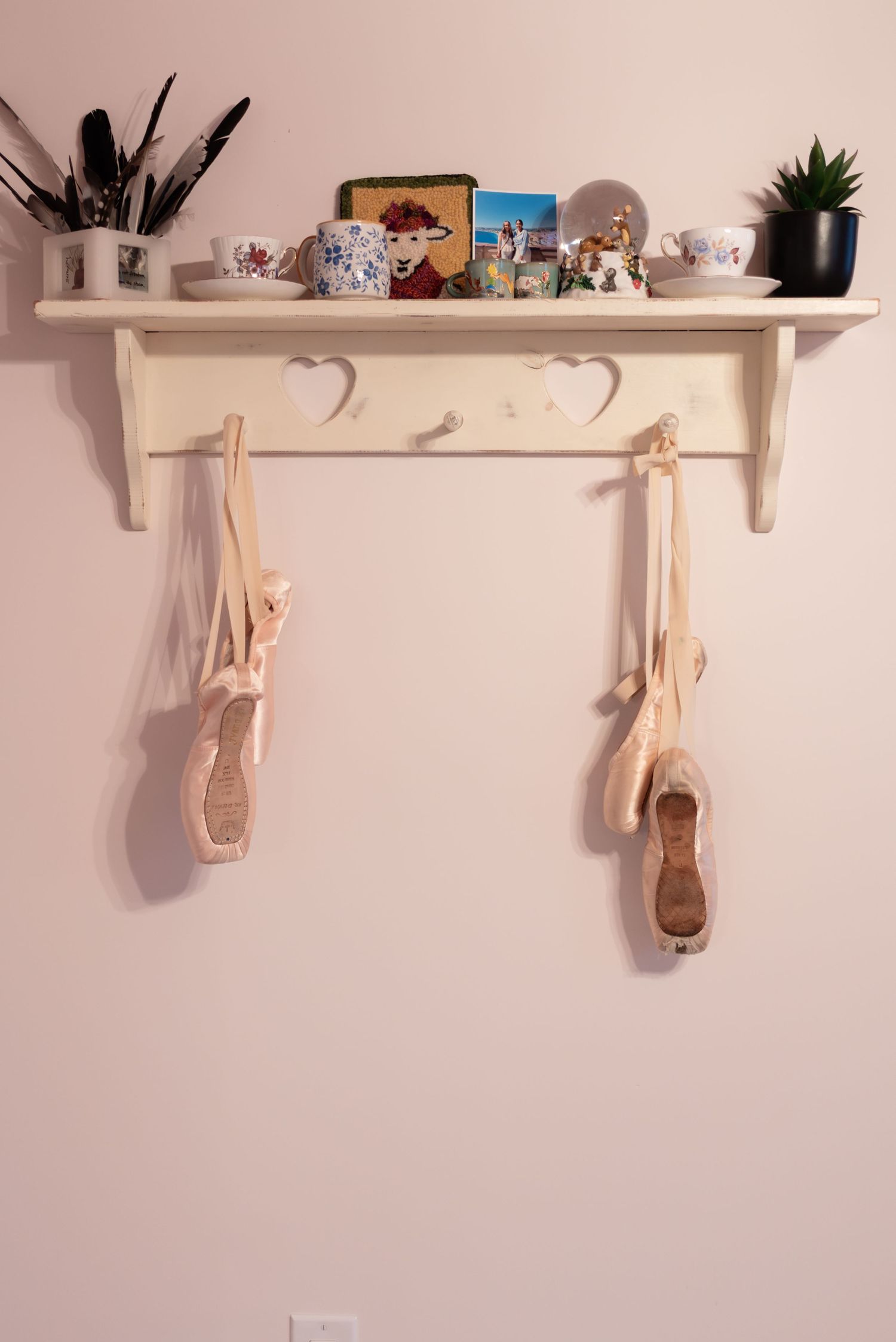 White wall shelf with heart hooks displaying ballet pointe shoes, mugs, and decorative items on pink wall.