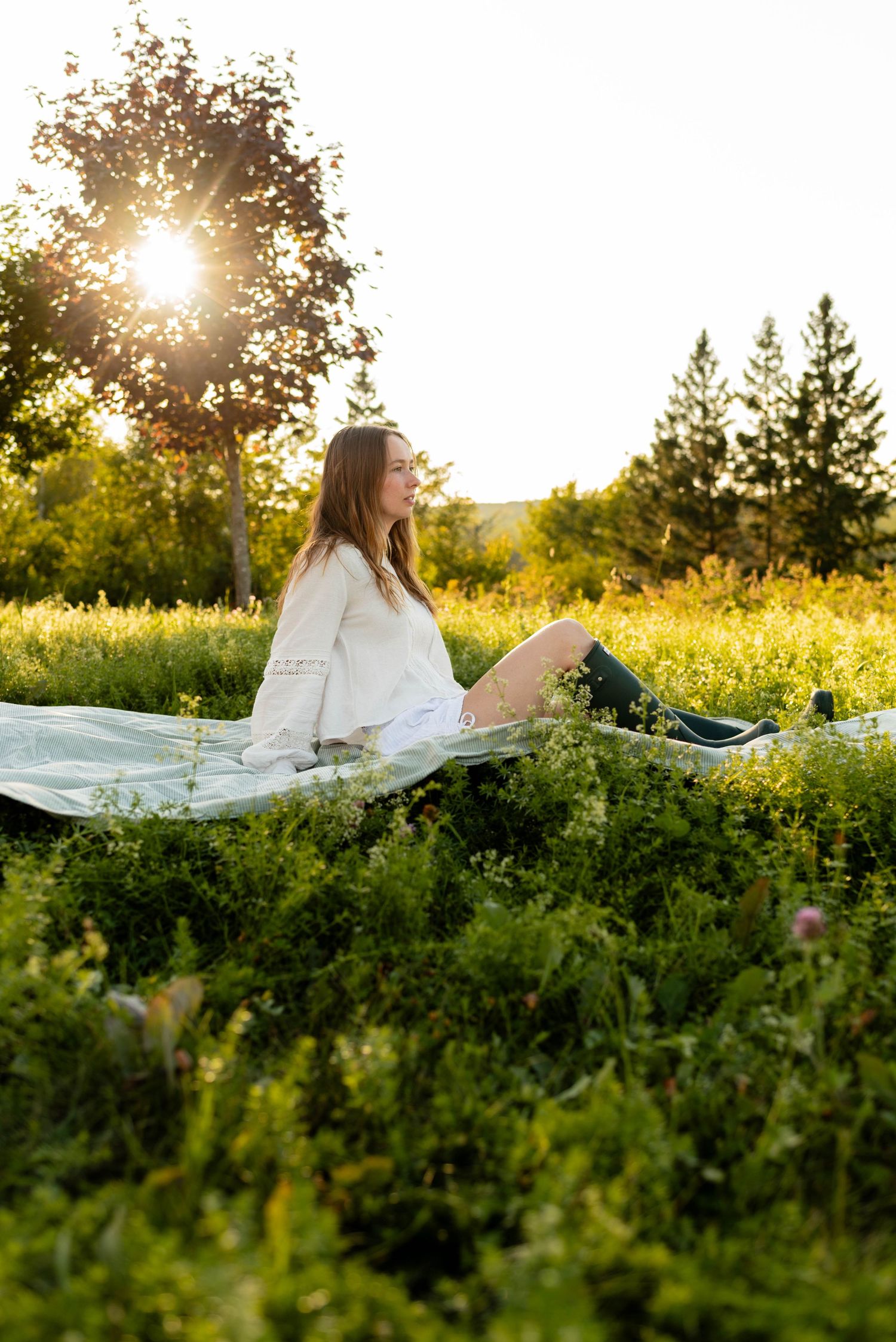 Woman in white top sitting on a blanket in a lush green meadow during golden sunset.