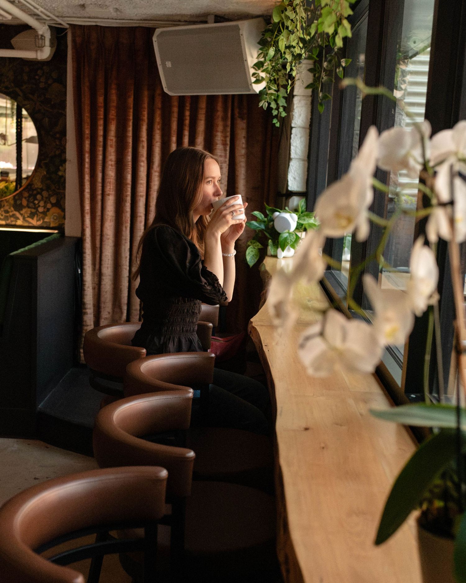 Woman enjoying a hot drink by sunlit cafe window surrounded by lush plants and white orchids.