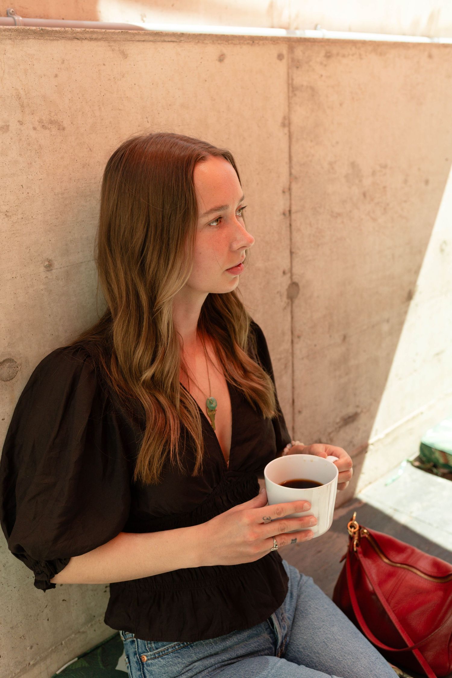 Young woman in black blouse holding a white coffee cup while leaning against a concrete wall outdoors.