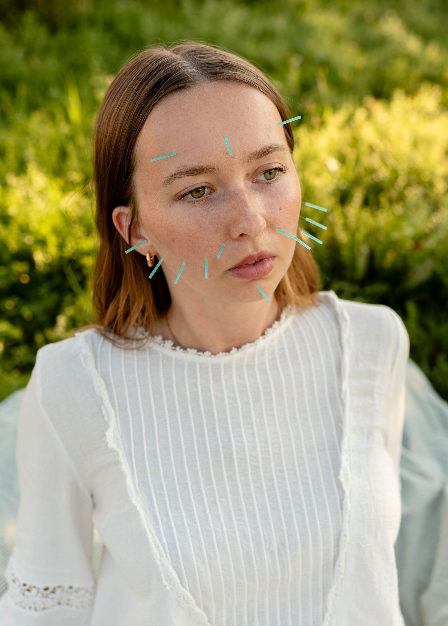 Woman in white top with acupuncture needles on face sitting outdoors in green garden.