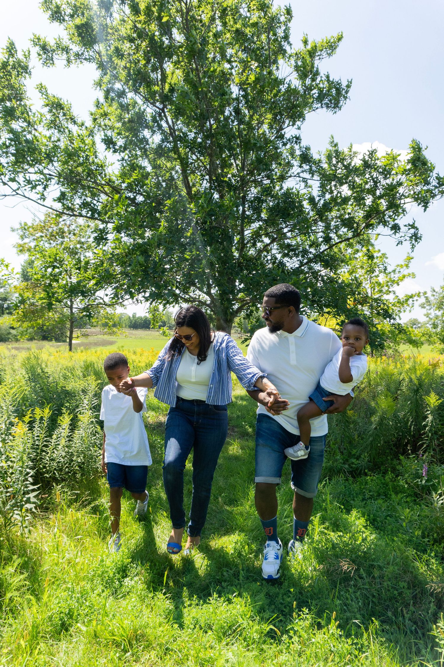 A joyful Black family of four walks through a lush green park on a sunny day, parents holding hands with their two young sons.