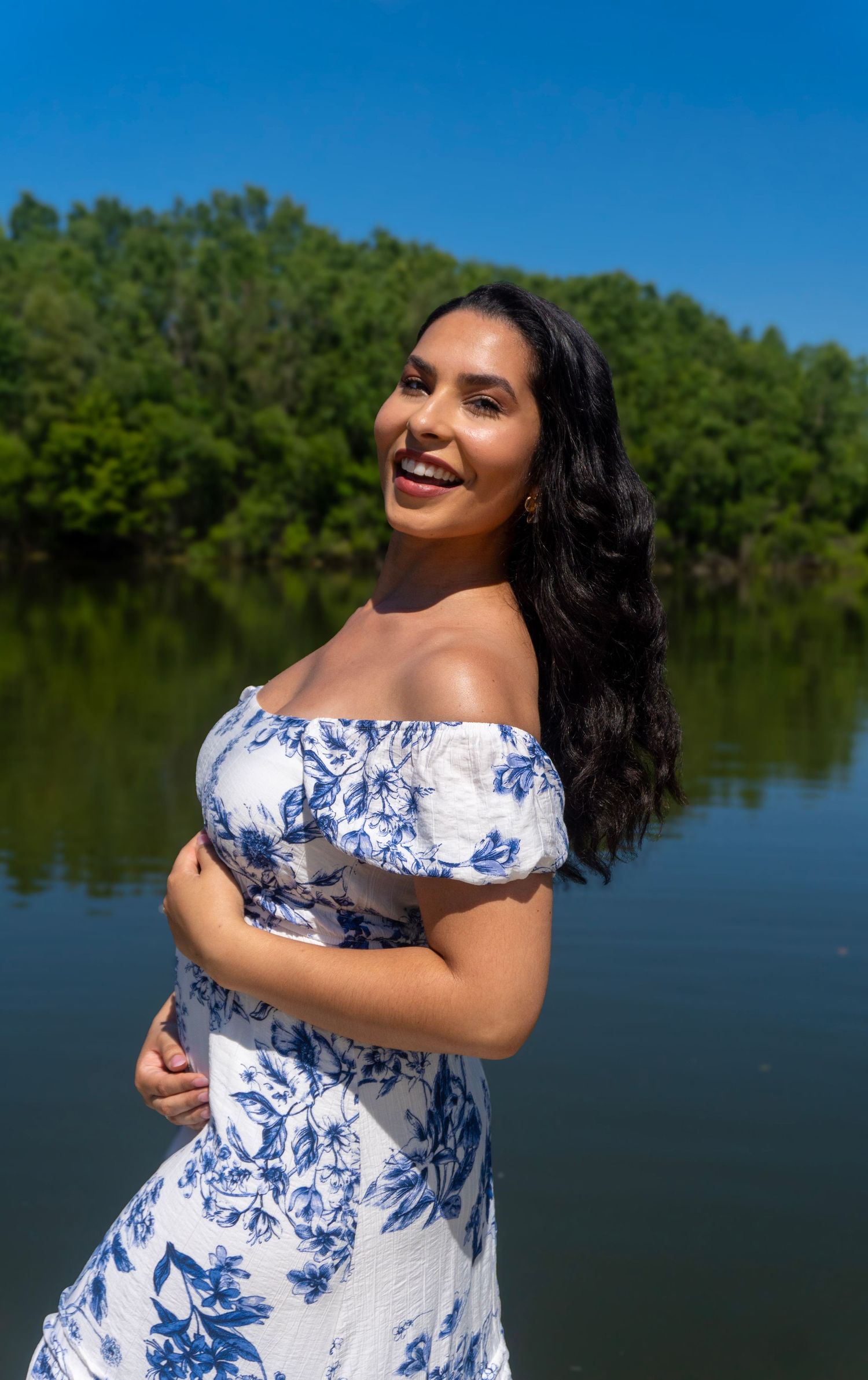 A smiling pregnant woman in a blue floral off-shoulder dress posing by a scenic lake outdoors.