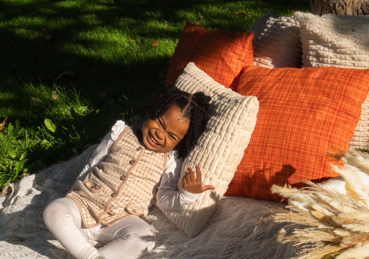 A young girl smiles while resting on cream and orange decorative pillows on a blanket outdoors in sunlight.
