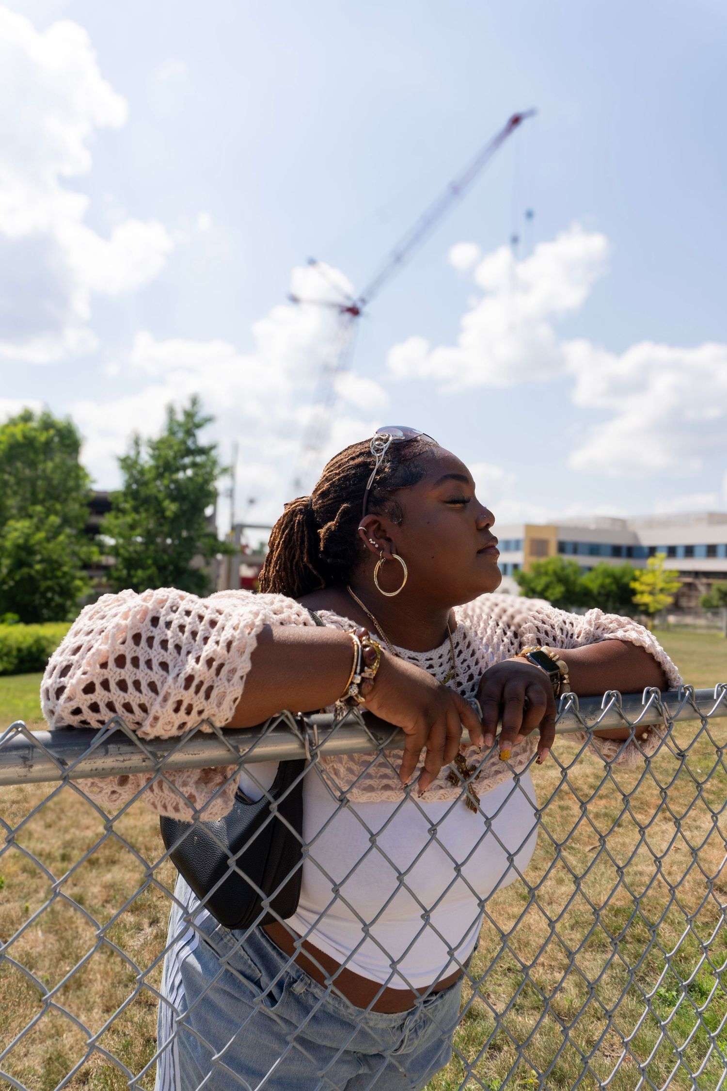 A young Black woman in a mesh cardigan leans on a chain-link fence with a construction crane in the background.