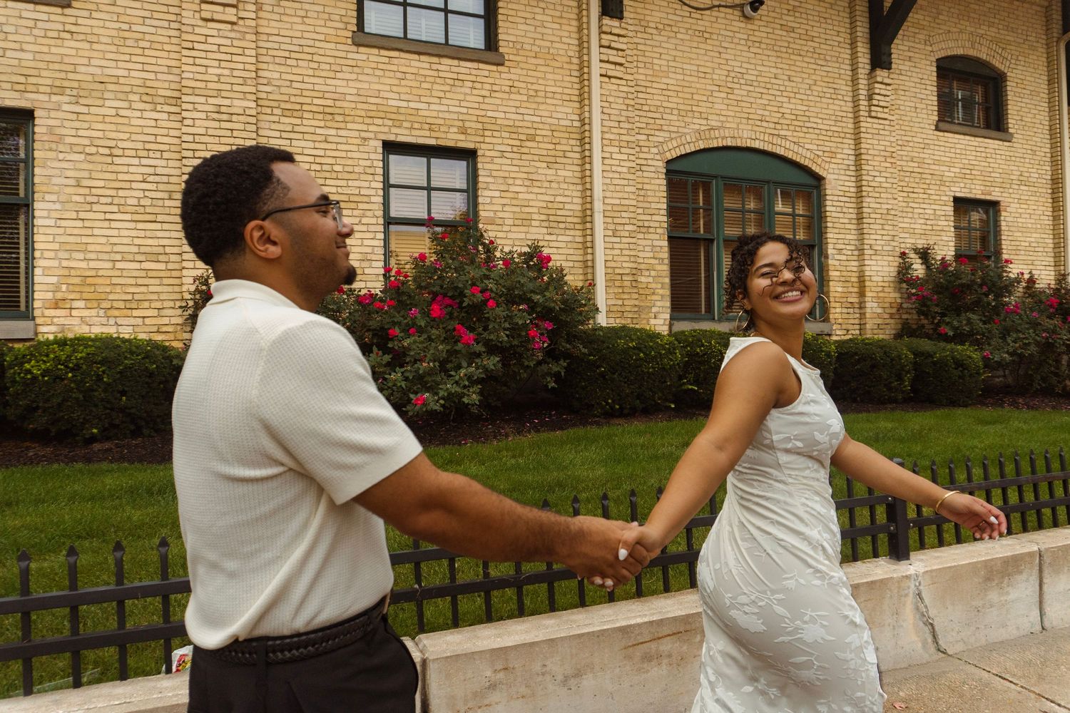A joyful couple in wedding attire holds hands and walks outside a brick building with lush greenery.