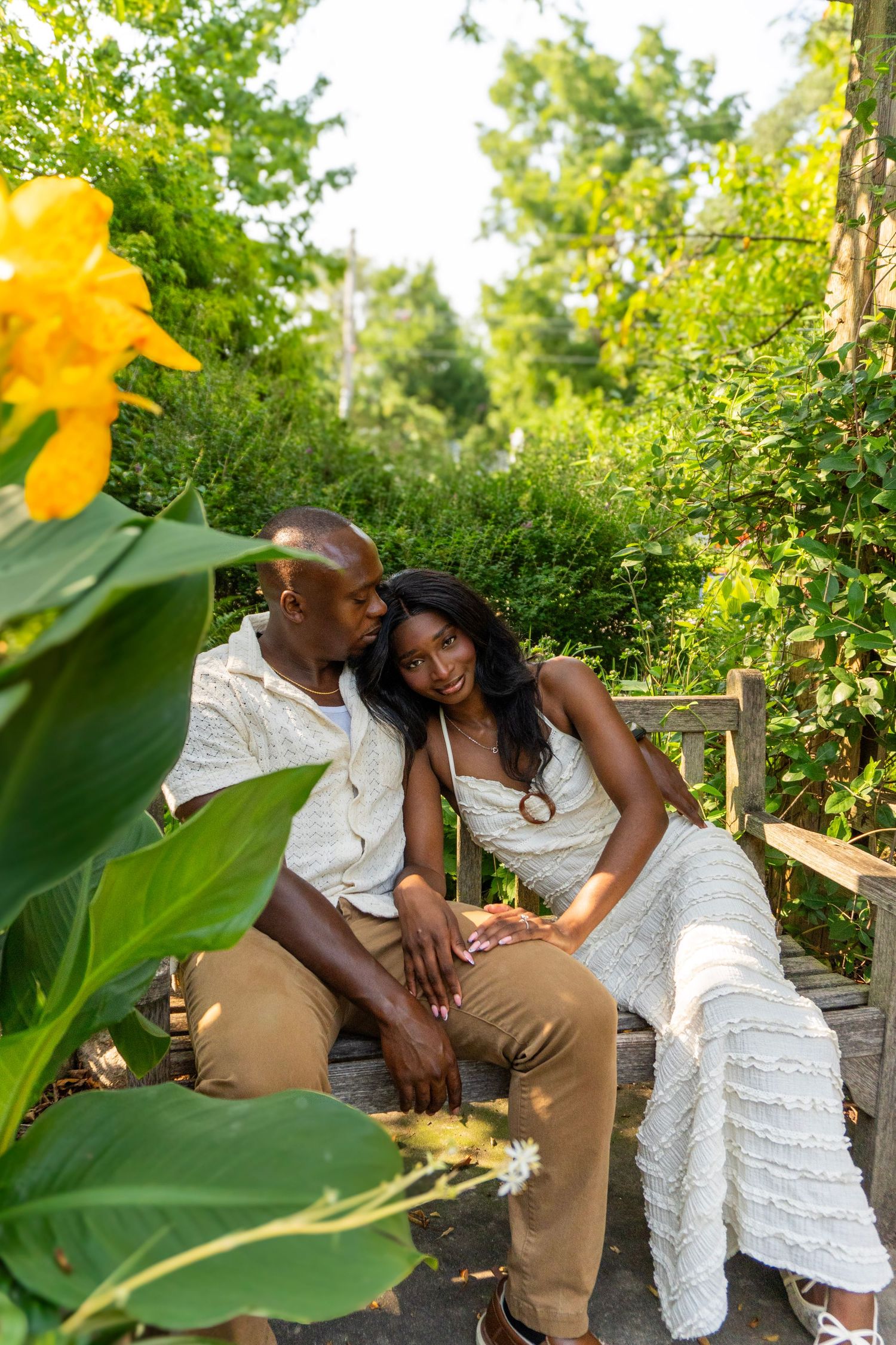 A couple sits on a bench in a lush garden, sharing a tender moment surrounded by green foliage and yellow flowers.