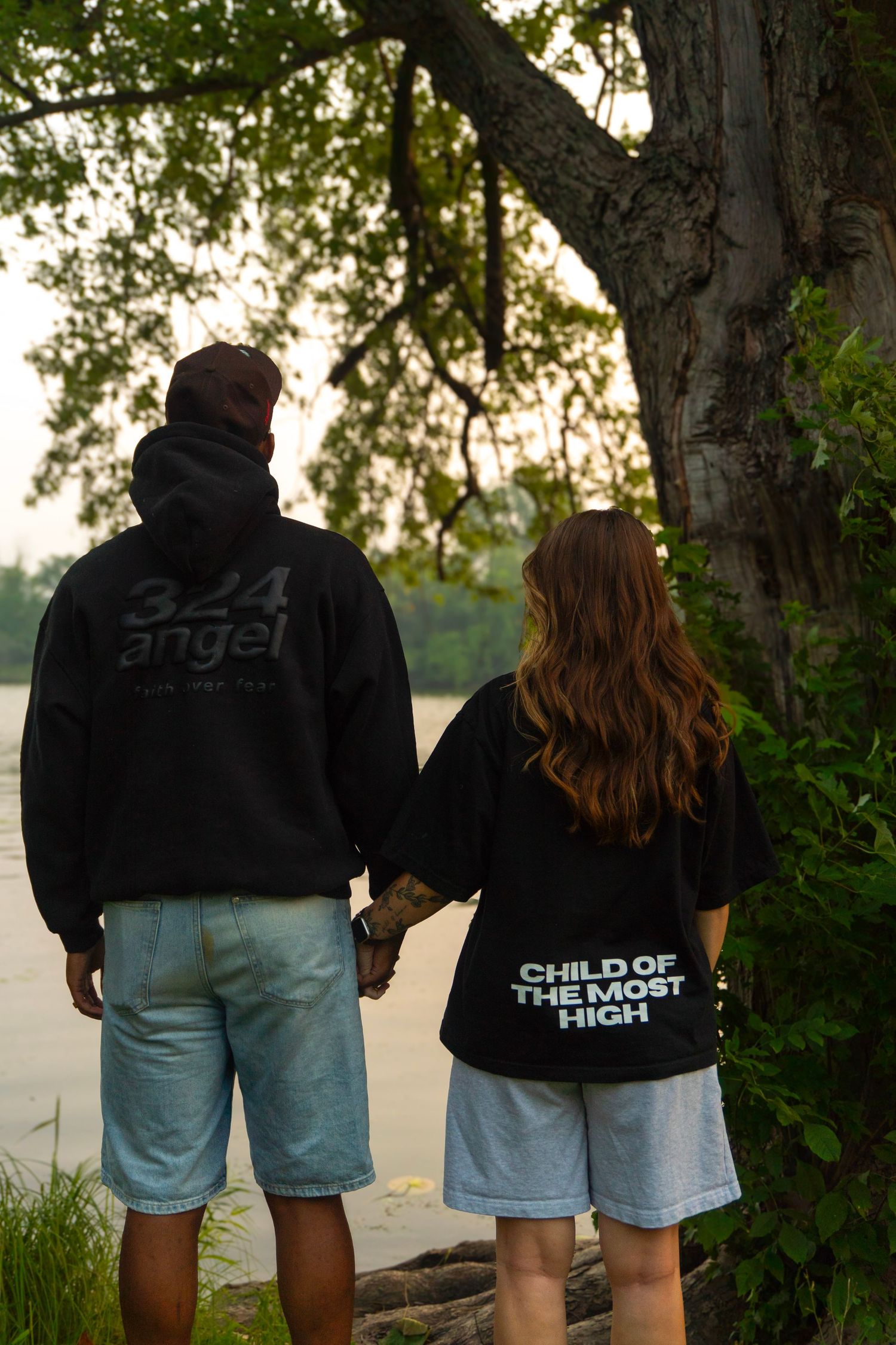 Couple holding hands from behind wearing graphic hoodies near tree by calm waterfront.