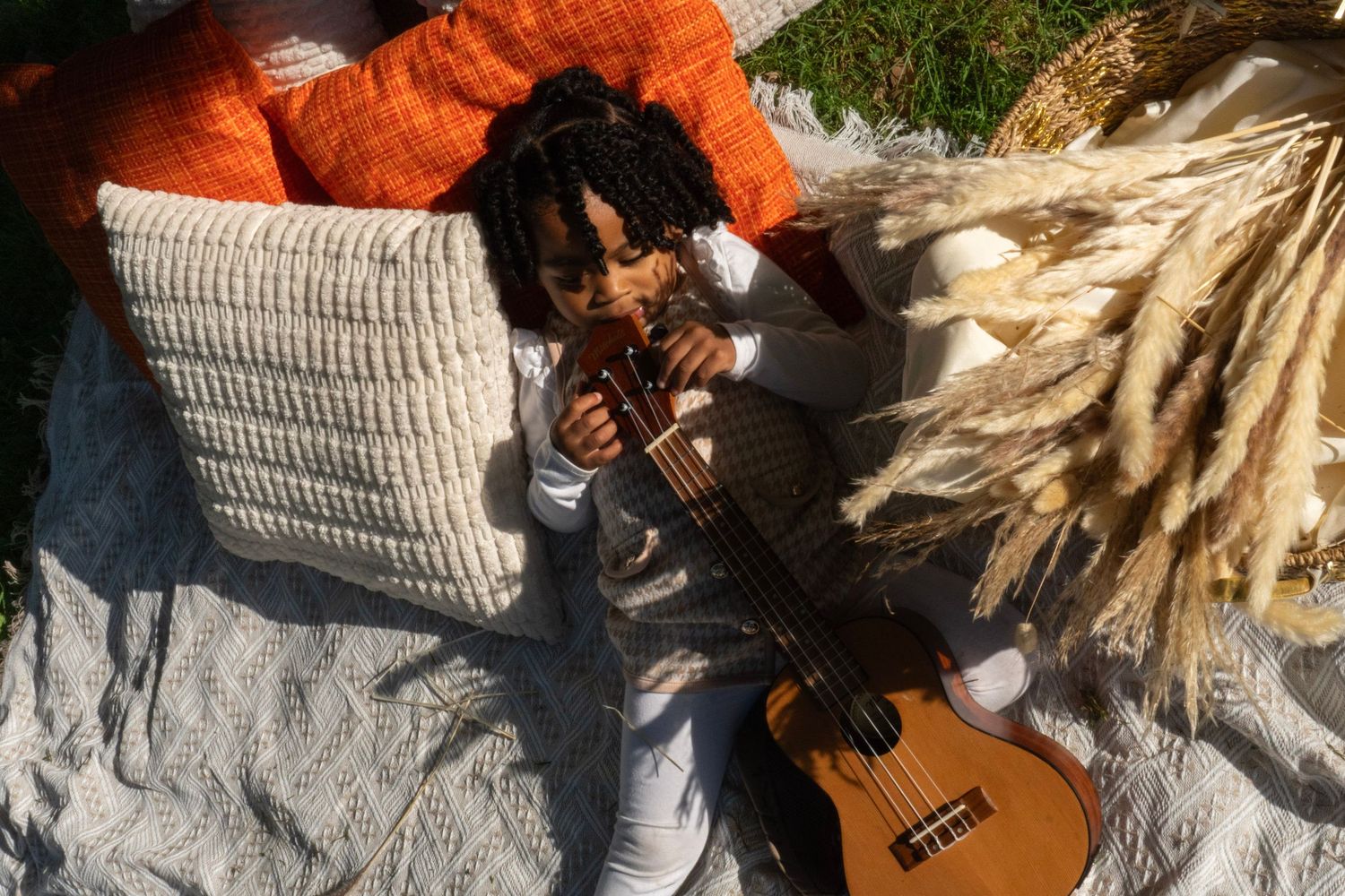 Young child with dreadlocks playing violin outdoors on blanket with ukulele nearby and dried pampas grass decor.