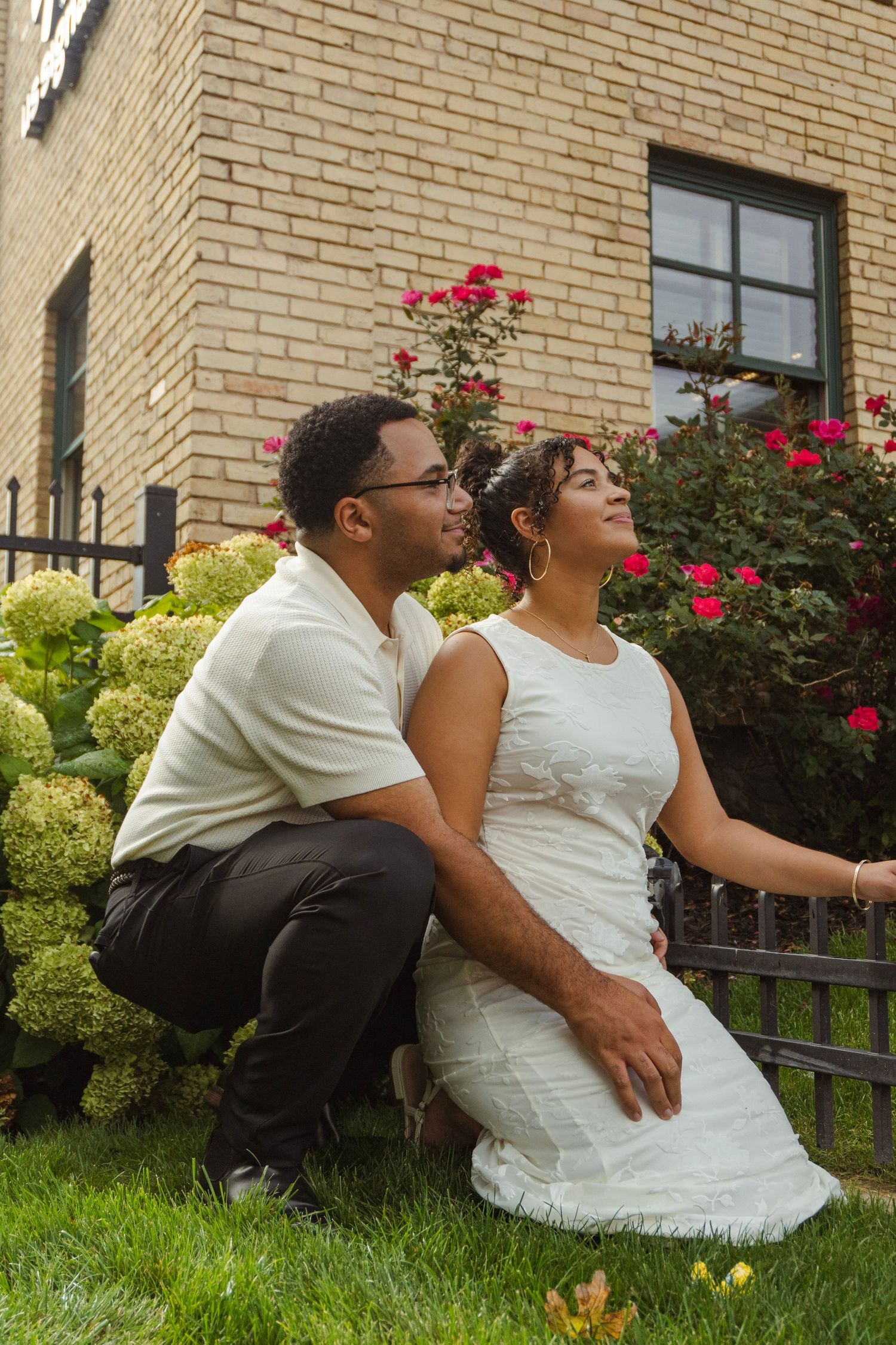A couple sits together on green grass in front of a brick building with roses, woman in white dress, man in white shirt.